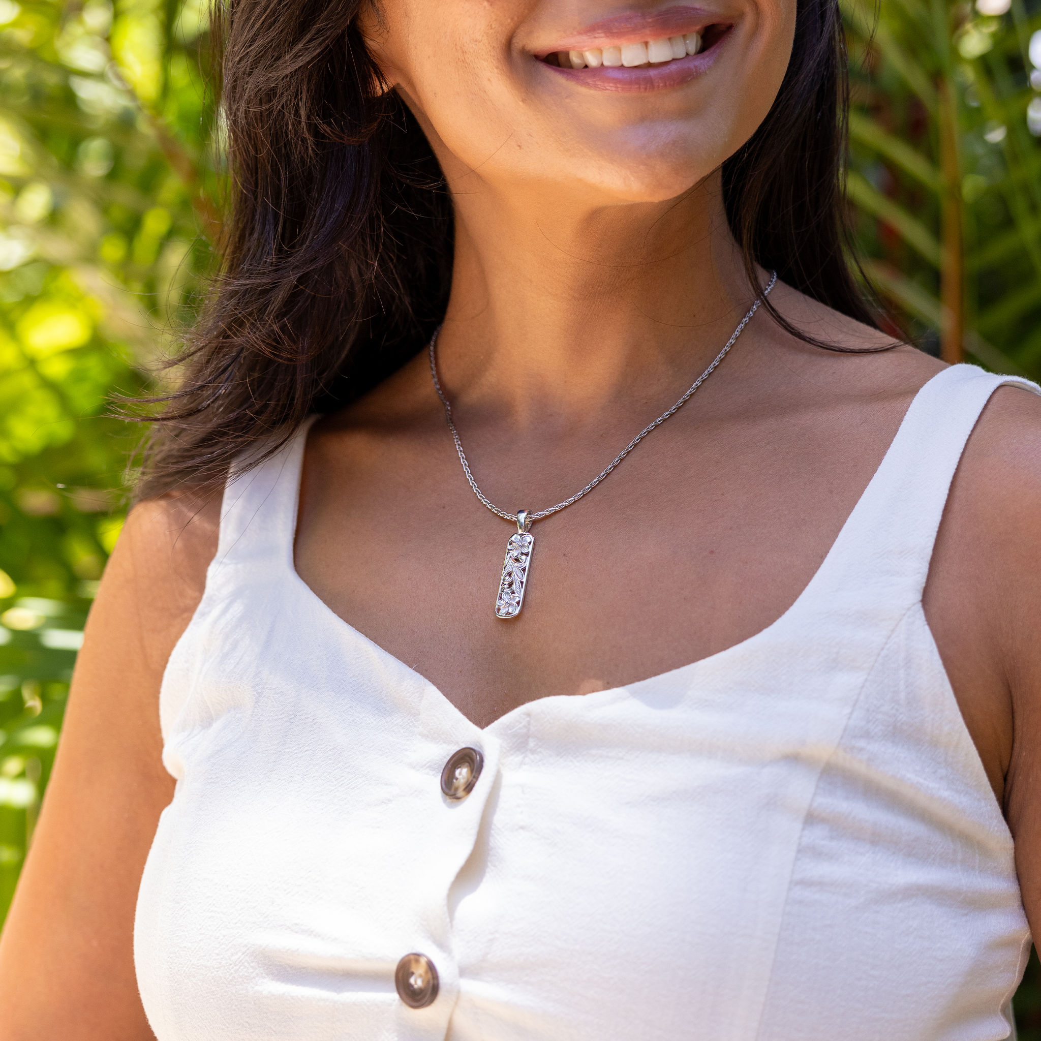 A woman wearing a sterling silver vertical rounded bar pendant necklace with white sapphire accented plumerias and maile leaf and fern scrollwork, photographed outdoors against a blurred green foliage background.