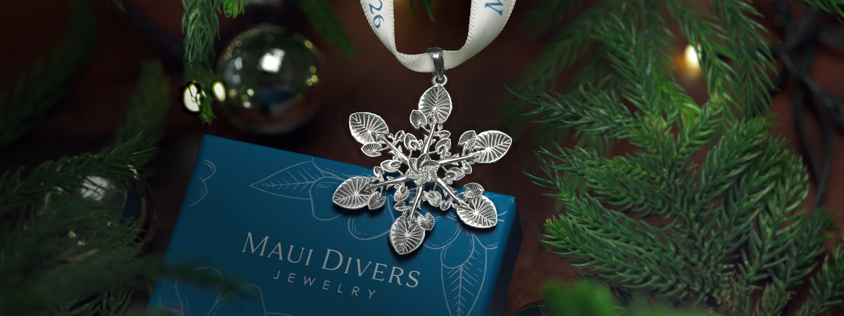 A silver Kalo leaf snowflake-style pendant ornament hanging from a white ribbon, displayed over a blue Maui Divers Jewelry box surrounded by green pine branches and holiday ornaments.