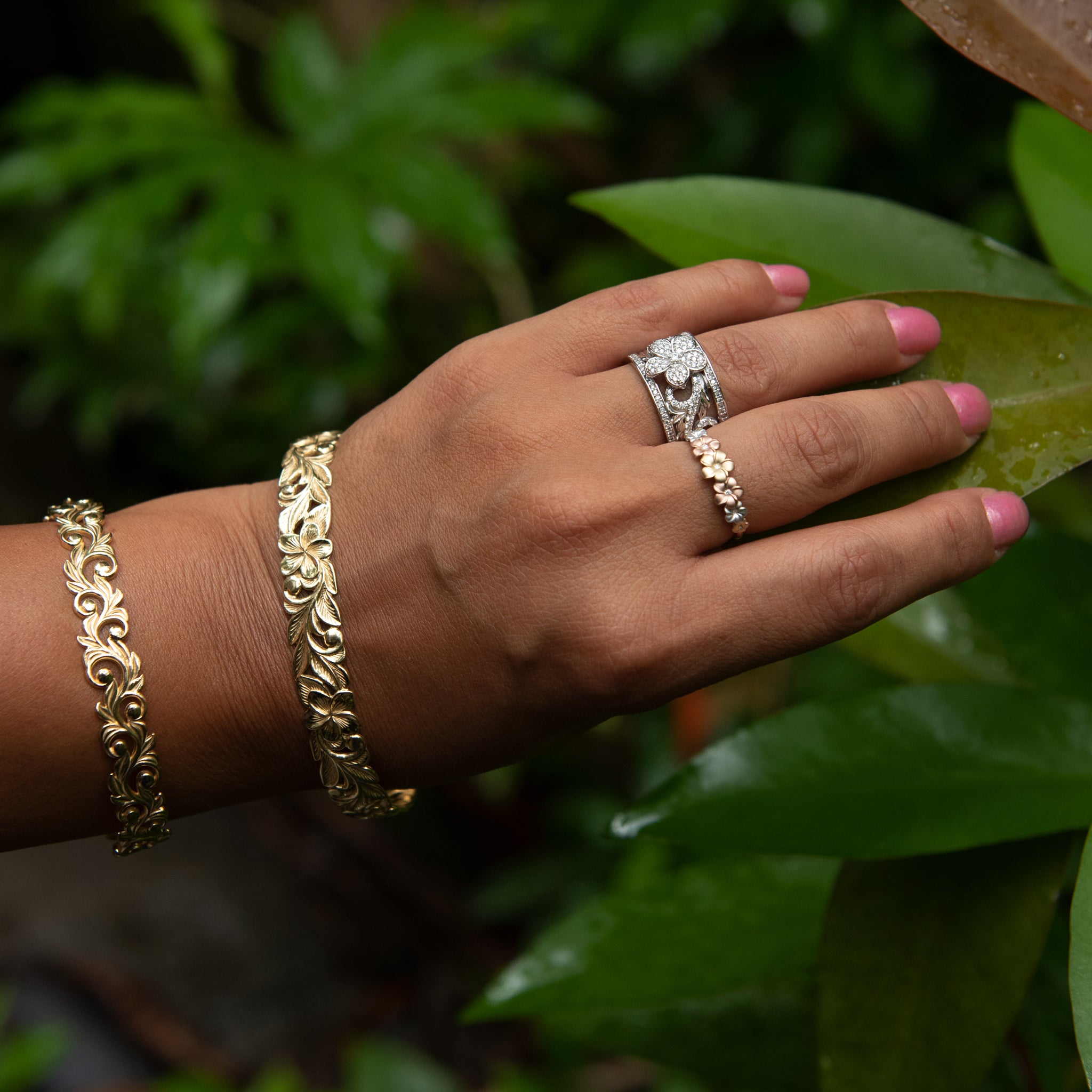 Hand wearing multiple Plumeria Rings in Gold with Diamonds and gold bracelets against a blurred leafy background