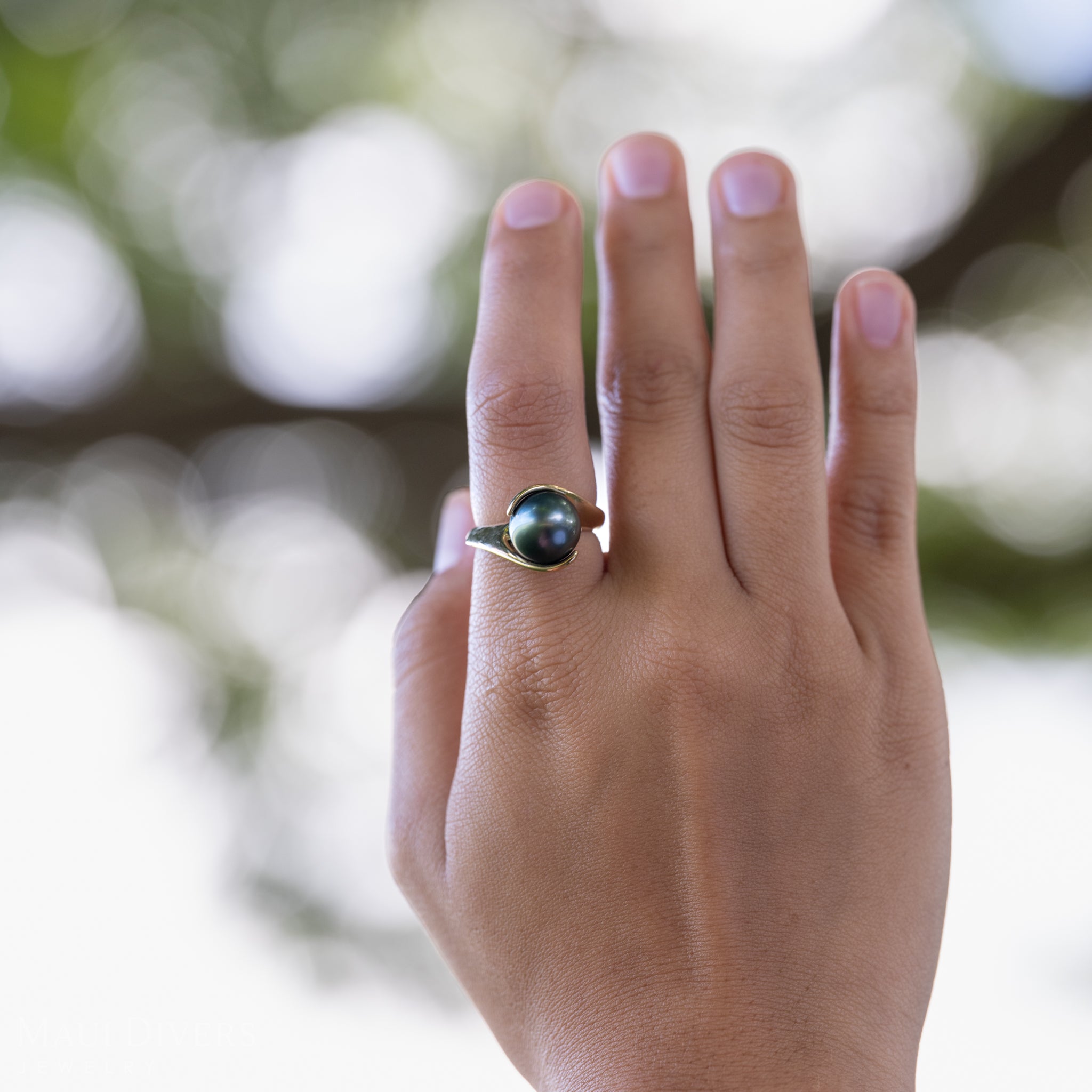 Close-up of a hand wearing a Tahitian Black Pearl Ring in 14k yellow gold on a pointer finger, against a blurred tree branch background