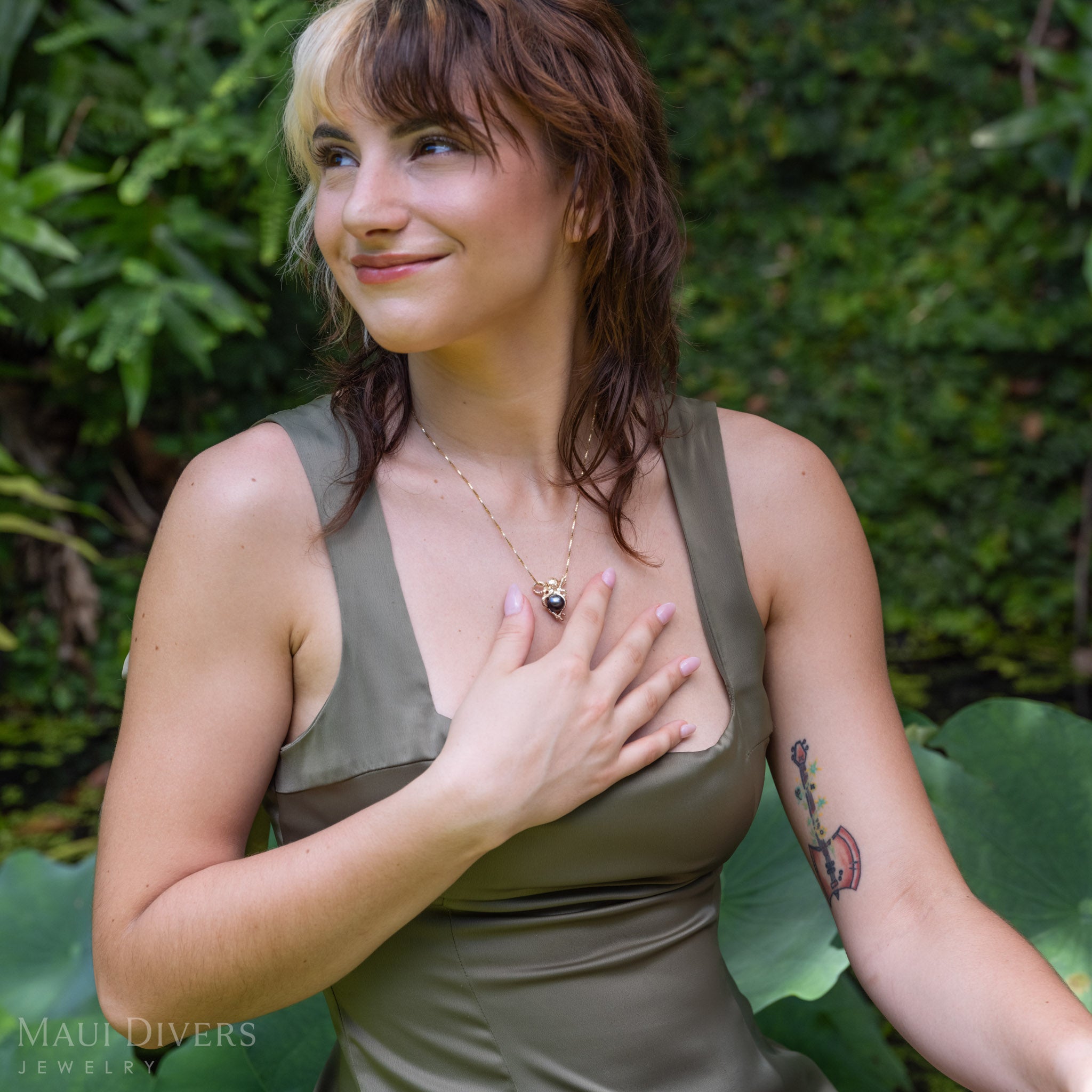 A lifestyle shot of a woman wearing a gold octopus pendant with a Tahitian black pearl wrapped in tentacles on a glistening box chain, styled with an olive green satin top against a blurry garden background.