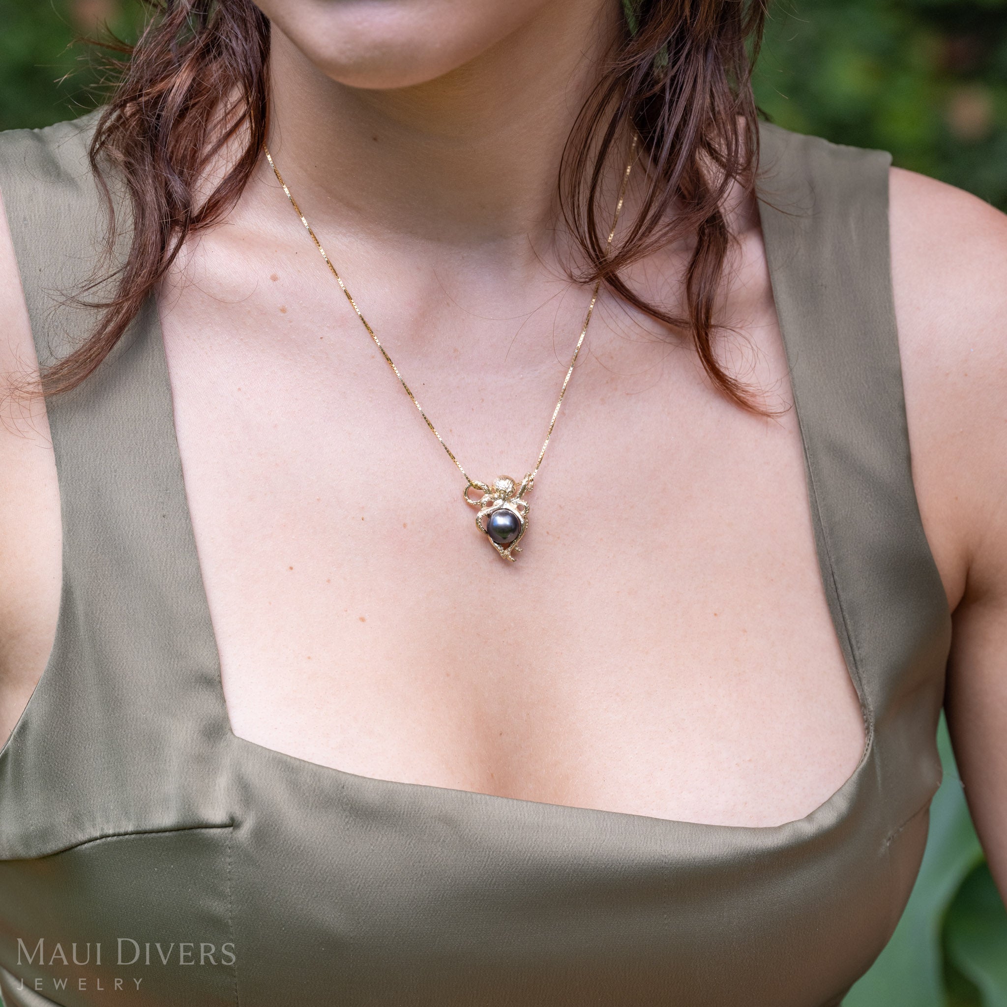 A close-up lifestyle shot of a woman wearing a gold octopus pendant with a Tahitian black pearl wrapped in tentacles on a glistening box chain, styled with an olive green satin top. 