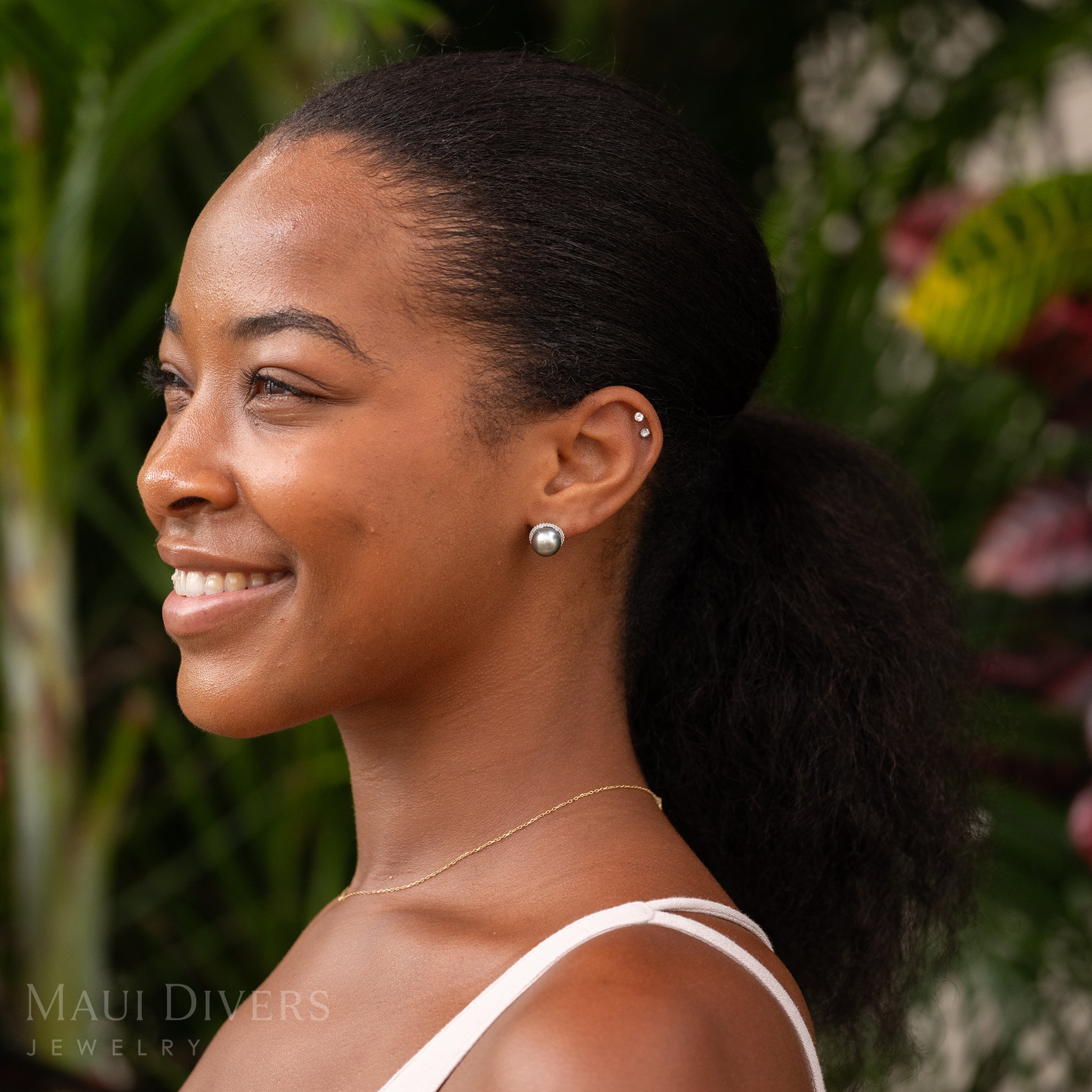A smiling woman wearing a Tahitian Black Pearl Diamond Halo Earrings in 14k White Gold on her left ear against a blurred tropical background