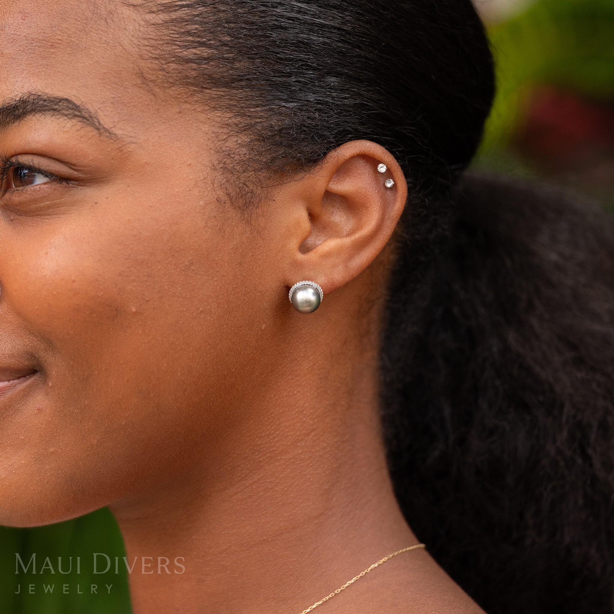 Close-up of a smiling woman wearing a Tahitian Black Pearl Diamond Halo Earrings in 14k White Gold on her left ear against a blurred background
