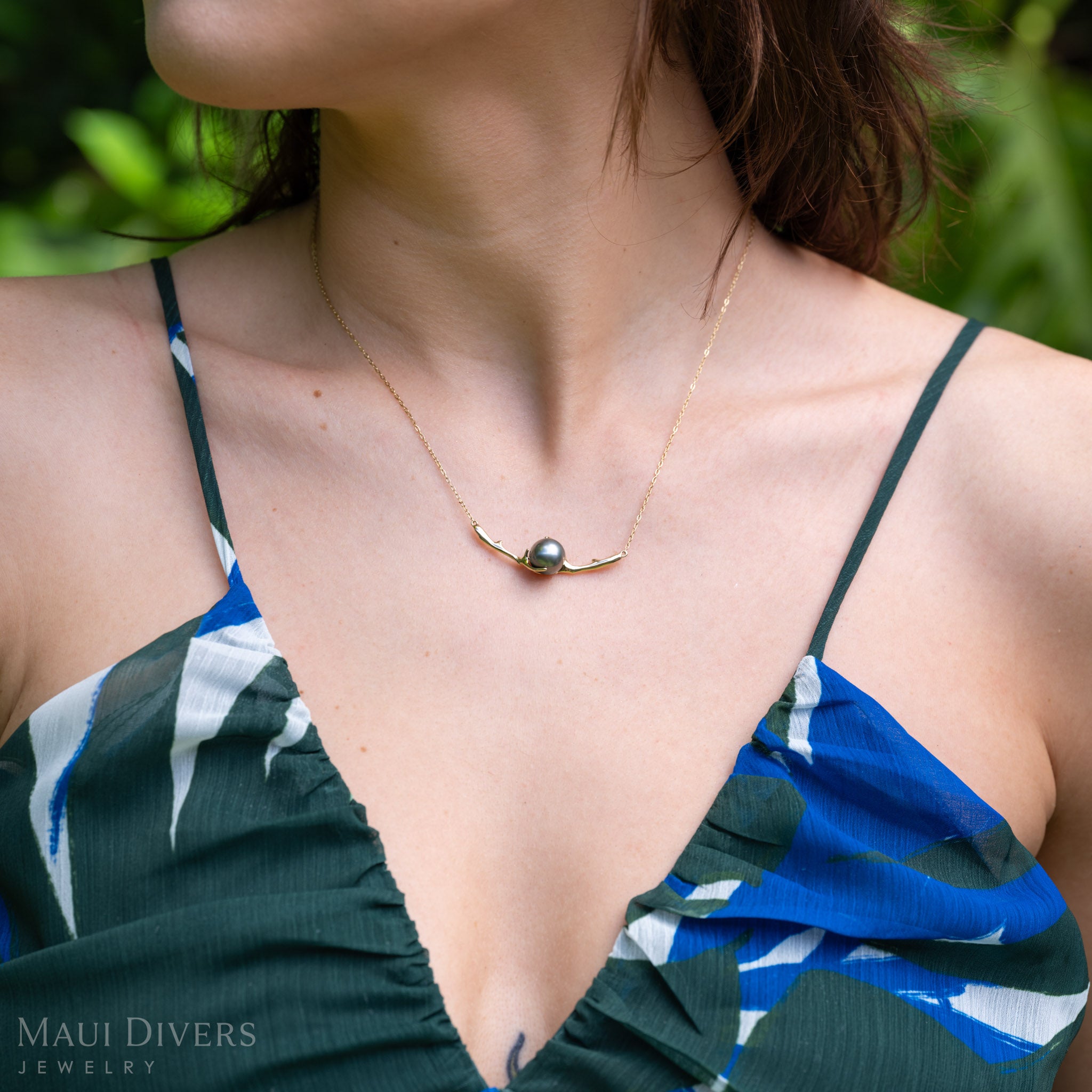 A close-up lifestyle shot of a model wearing a gold necklace with a curved heritage collection black coral-inspired design with a Tahitian black pearl in the middle, paired with a blue and green patterned top.