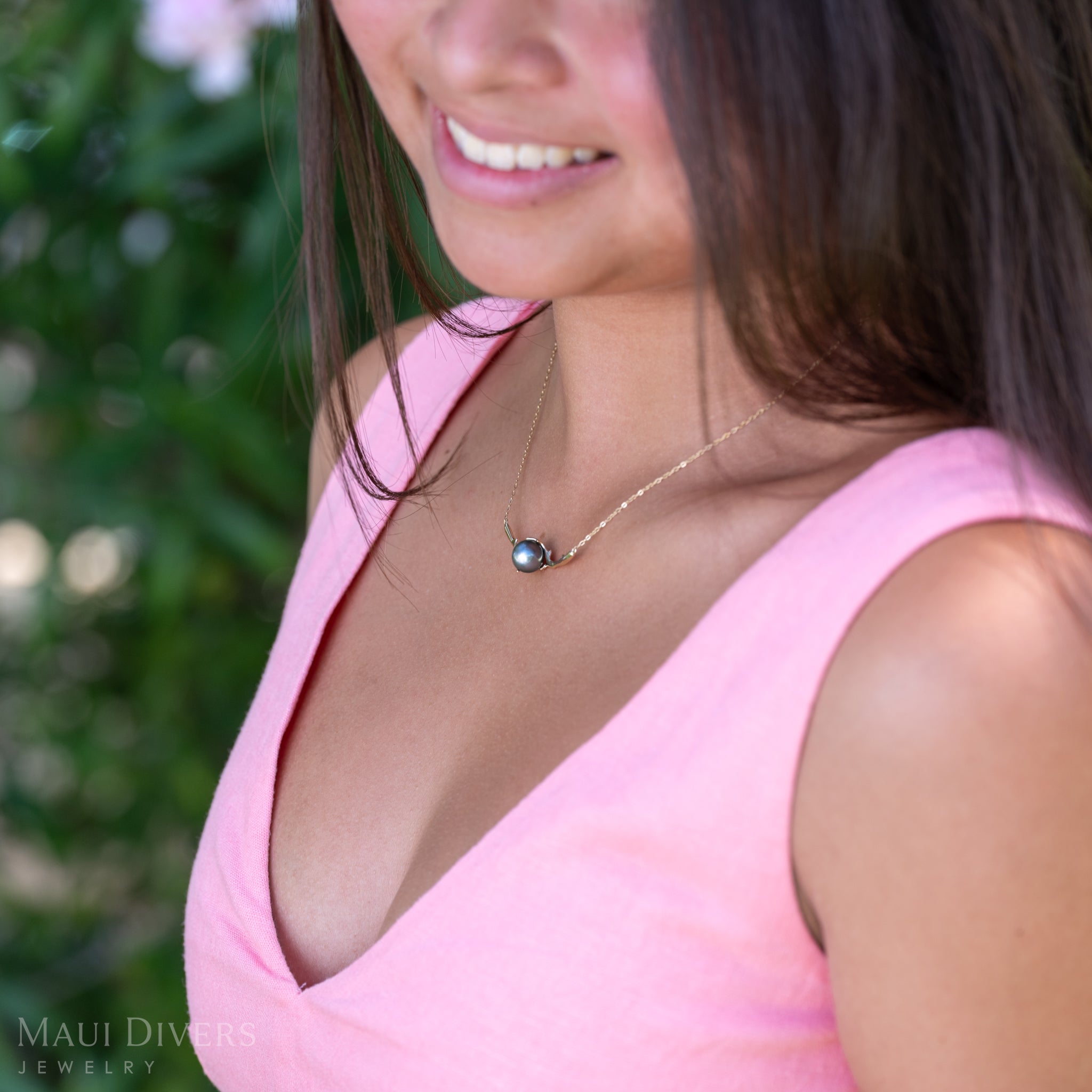 A close-up of a smiling woman in a light pink top wearing a Heritage Tahitian Black Pearl Necklace in 14k yellow gold against a blurred leafy background