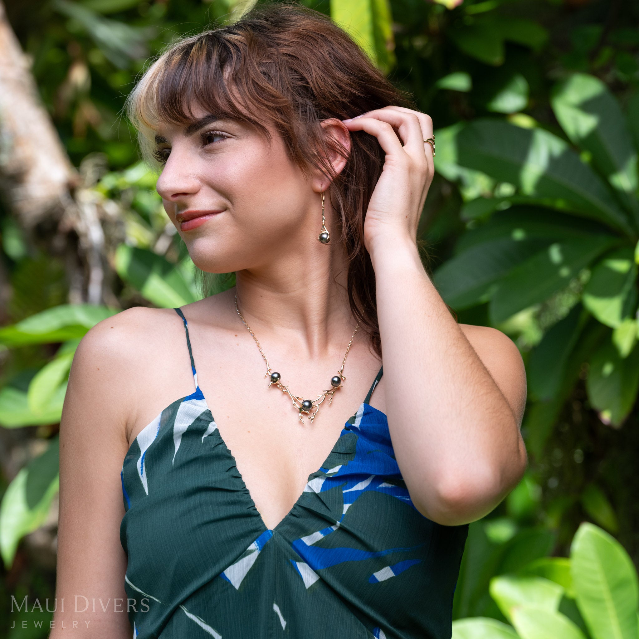 A lifestyle shot of a model wearing a Tahitian black pearl necklace with black coral-branch inspired designs in 14k yellow gold, along with matching dangling earrings. The model is wearing a blue and green patterned top against a green foliage background.