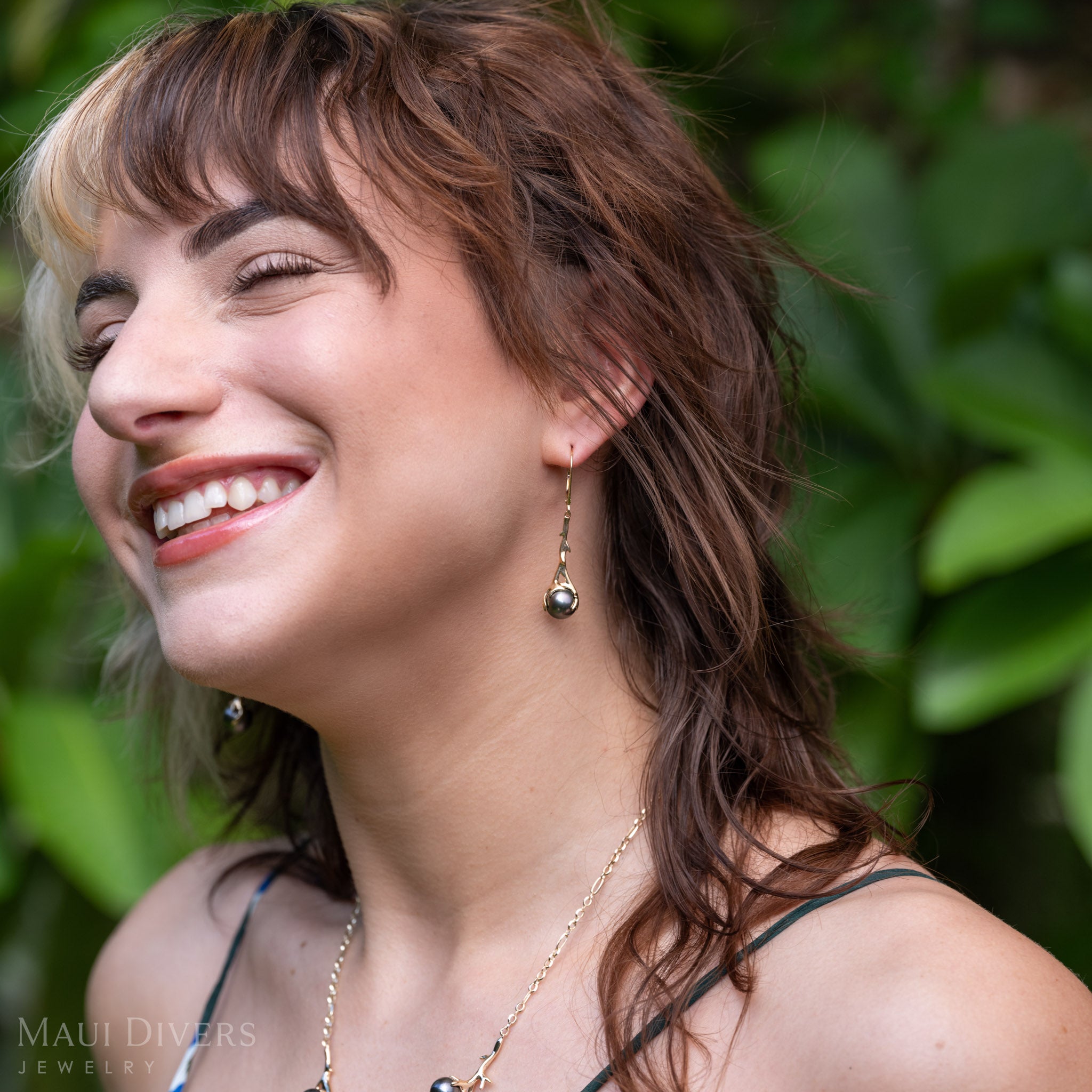 A lifestyle photo of a woman wearing a Heritage Collection black Coral-branch inspired leverback earrings in solid 14k yellow gold with a Tahitian black pearl against a blurry leafy background