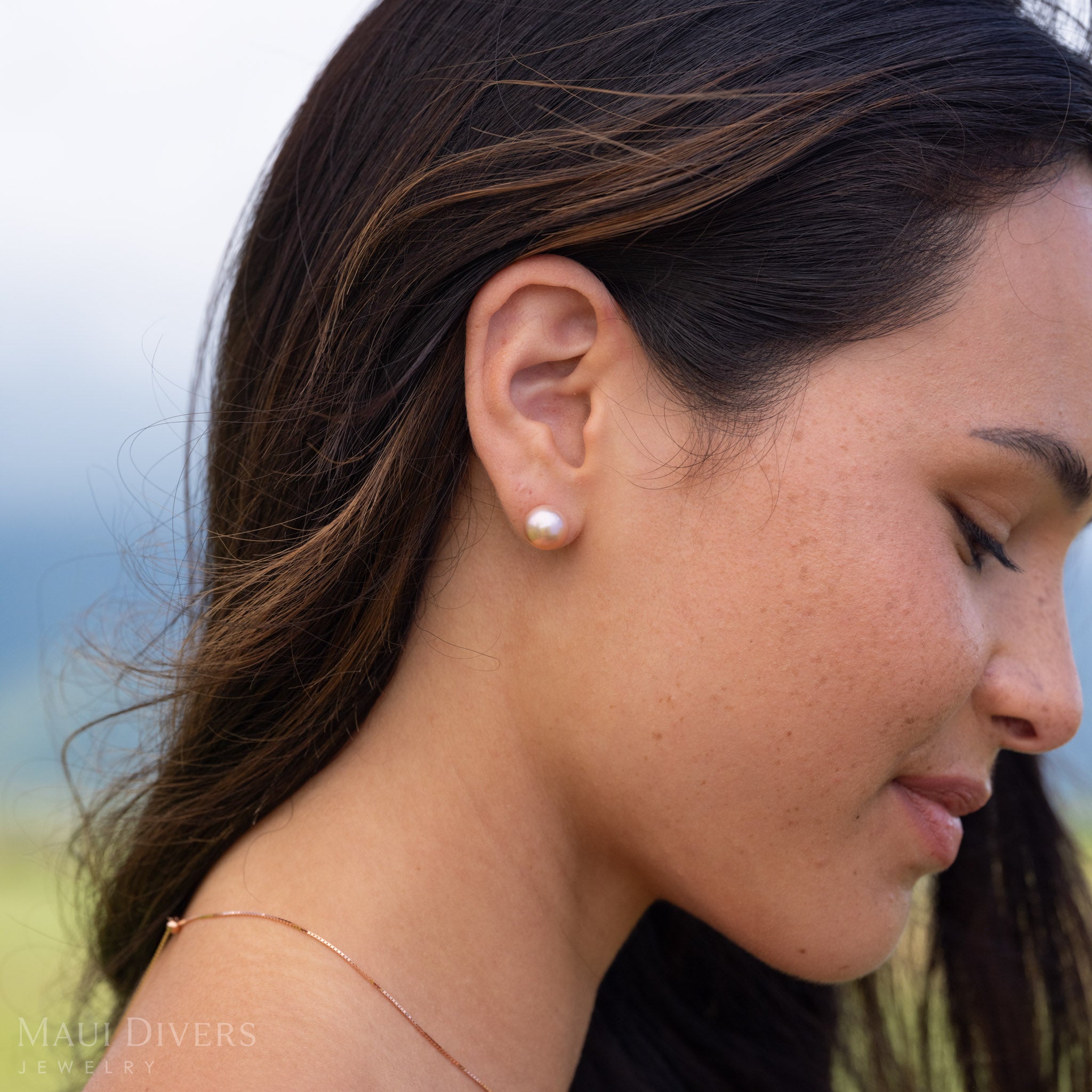 Close-up of a woman wearing a freshwater pearl natural color stud earring in 14k yellow gold with a blurry green and blue background