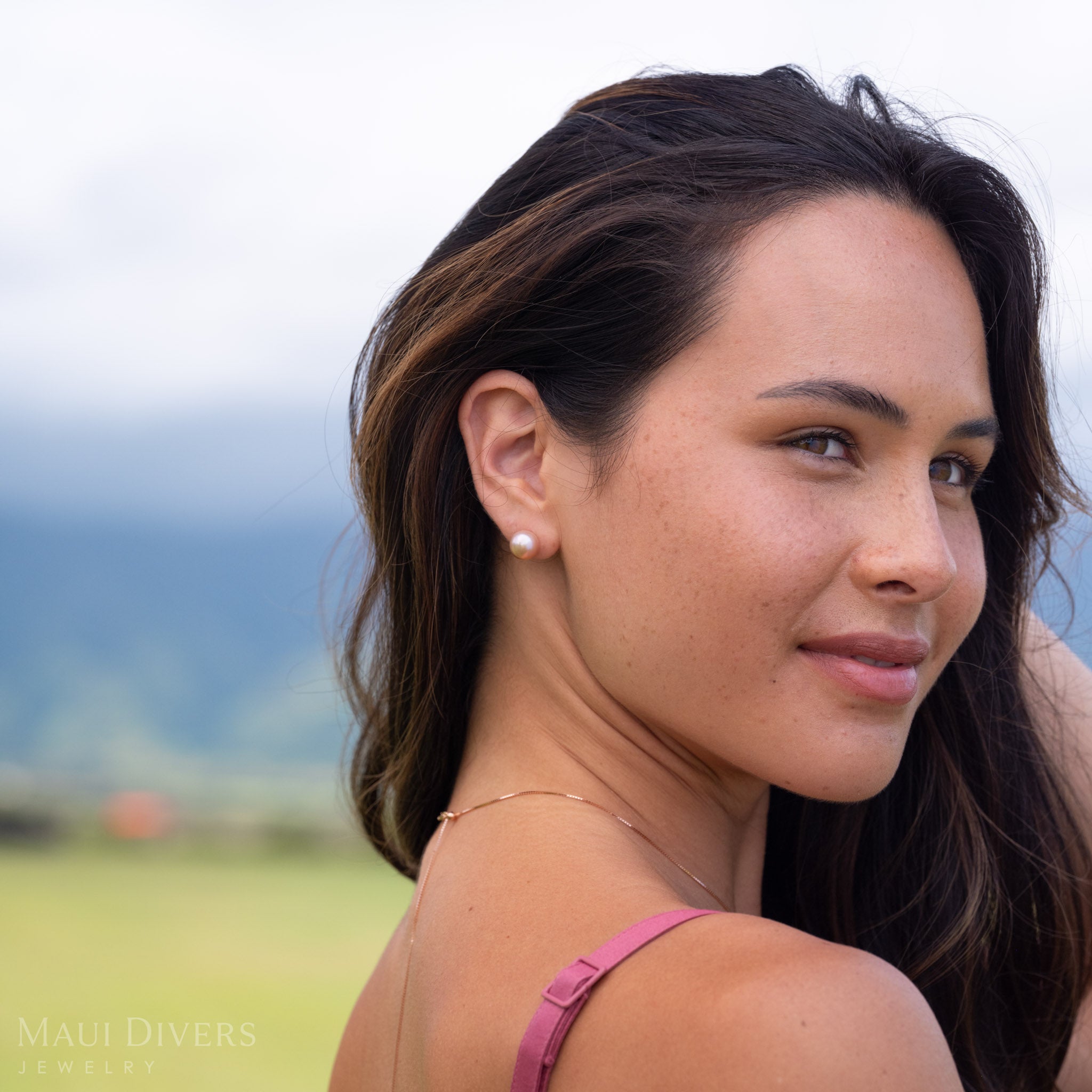 A woman looking over her shoulder wearing a freshwater pearl natural color stud earring in 14k yellow gold with a blurry green and blue ocean background