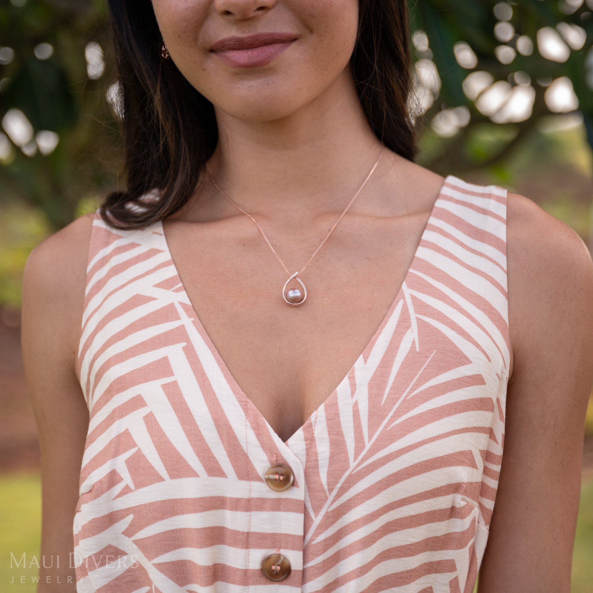 A woman wearing a teardrop-shaped Lilac Freshwater Pearl Pendant in 14k Rose Gold with a pink and white top and blurred green trees in the background.