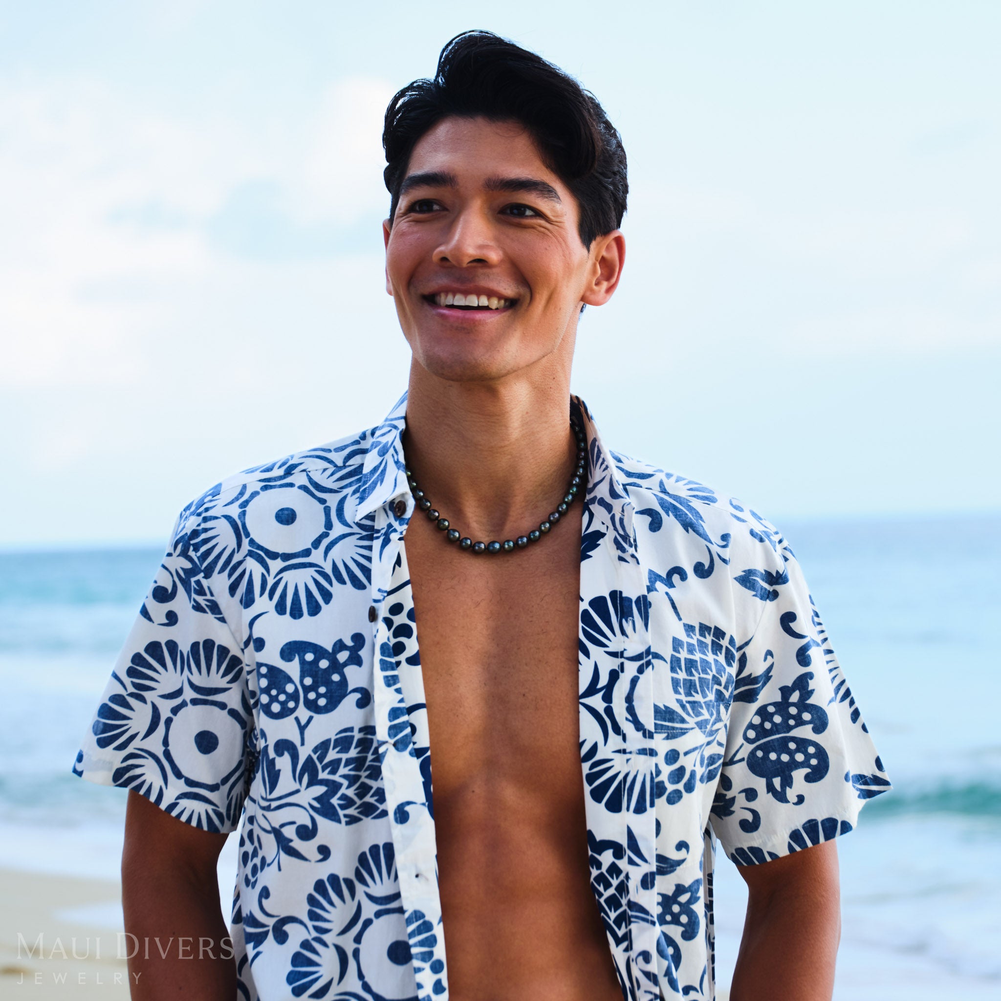 Man wearing a blue and white shirt with a Tahitian Black Pearl Strand against a blurred beach background