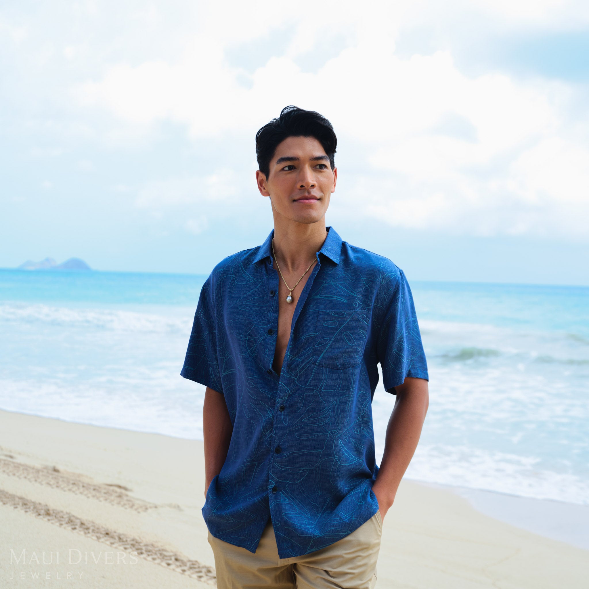 Man wearing a blue patterned shirt with a Waterfall Tahitian Black Pearl Pendant in Gold against a blurred beach background