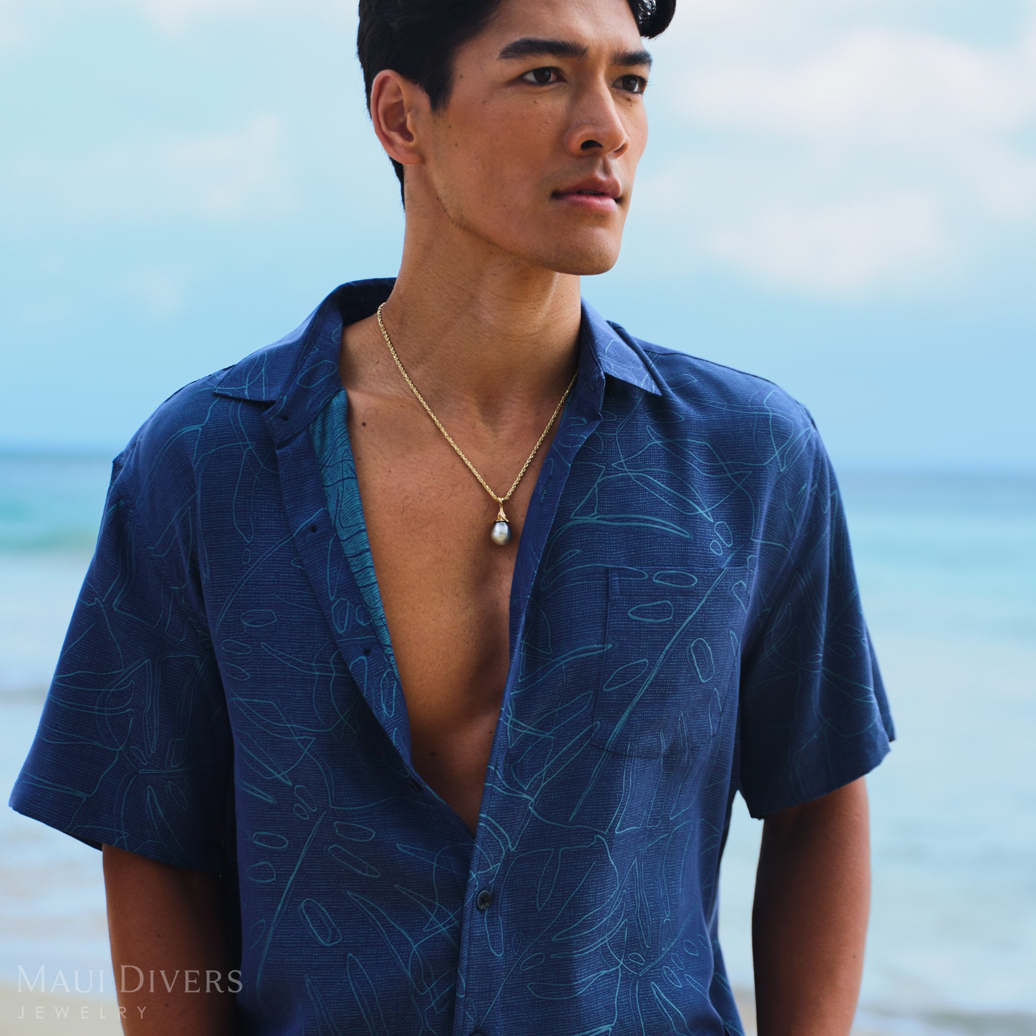 Man wearing a blue patterned shirt with a Waterfall Tahitian Black Pearl Pendant in Gold against a blurred beach background