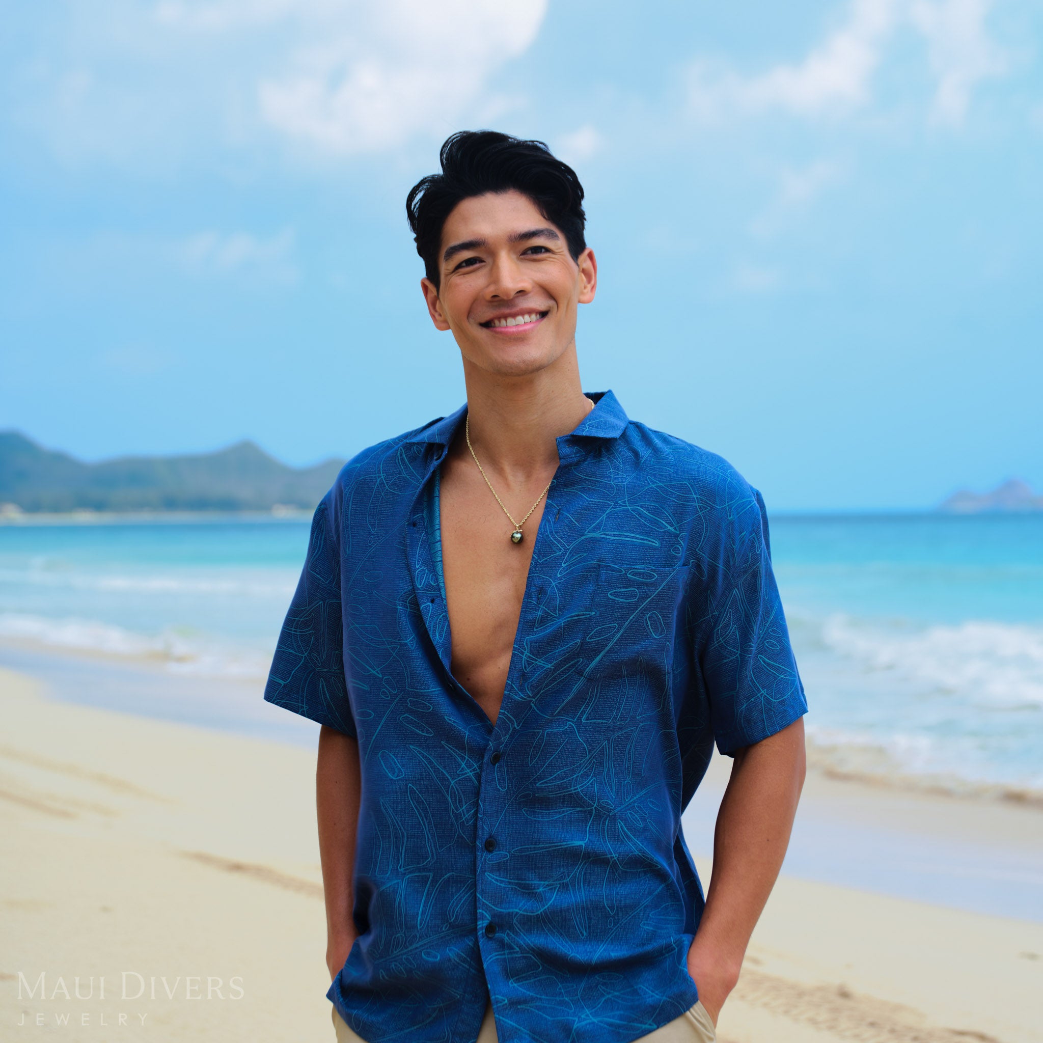 Man in a blue shirt  and wearing a gHonu 13-14mm Tahitian Black Pearl Pendant in Gold standing on a beach