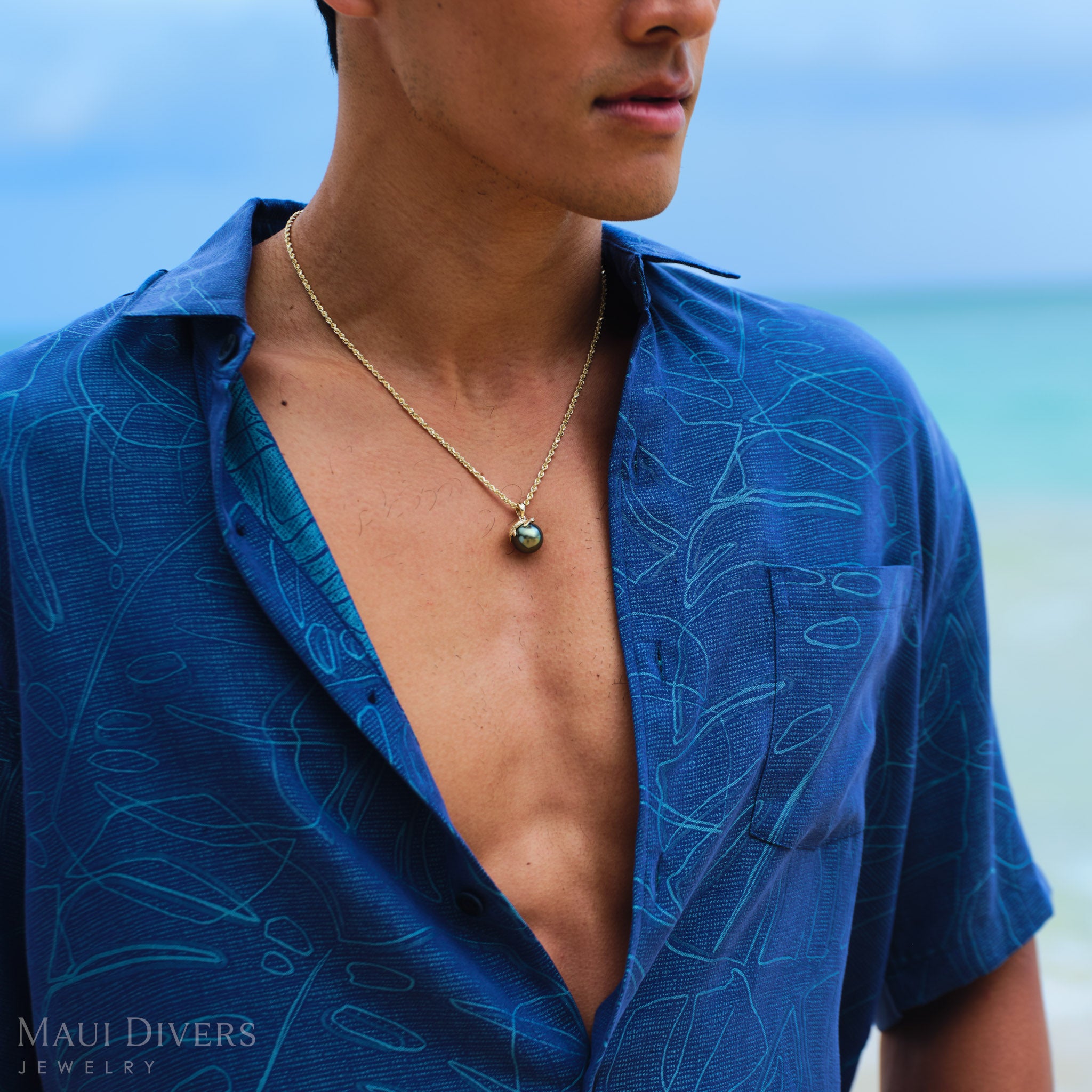 Man wearing a blue patterned shirt with a Honu 13-14mm Tahitian Black Pearl Pendant in Gold on a beach