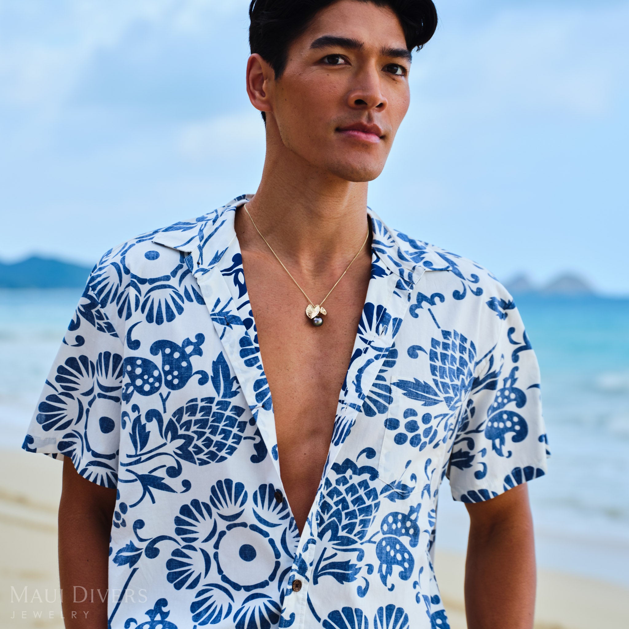 Man wearing a blue and white patterned shirt with a Kalo 12-15mm Circle Tahitian Black Pearl Pendant in Gold on a beach