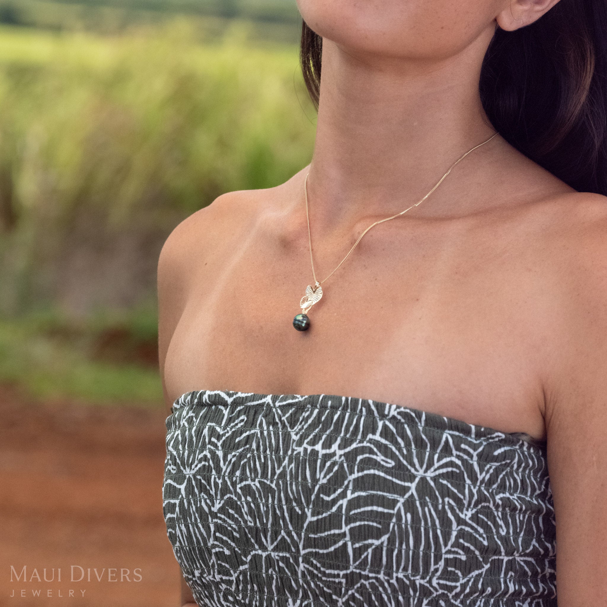 Side-angle close-up of a 14k Gold Kalo (Taro) Pendent with a Black Tahitian Pearl in front of a blurred green grass and brown dirt background. 