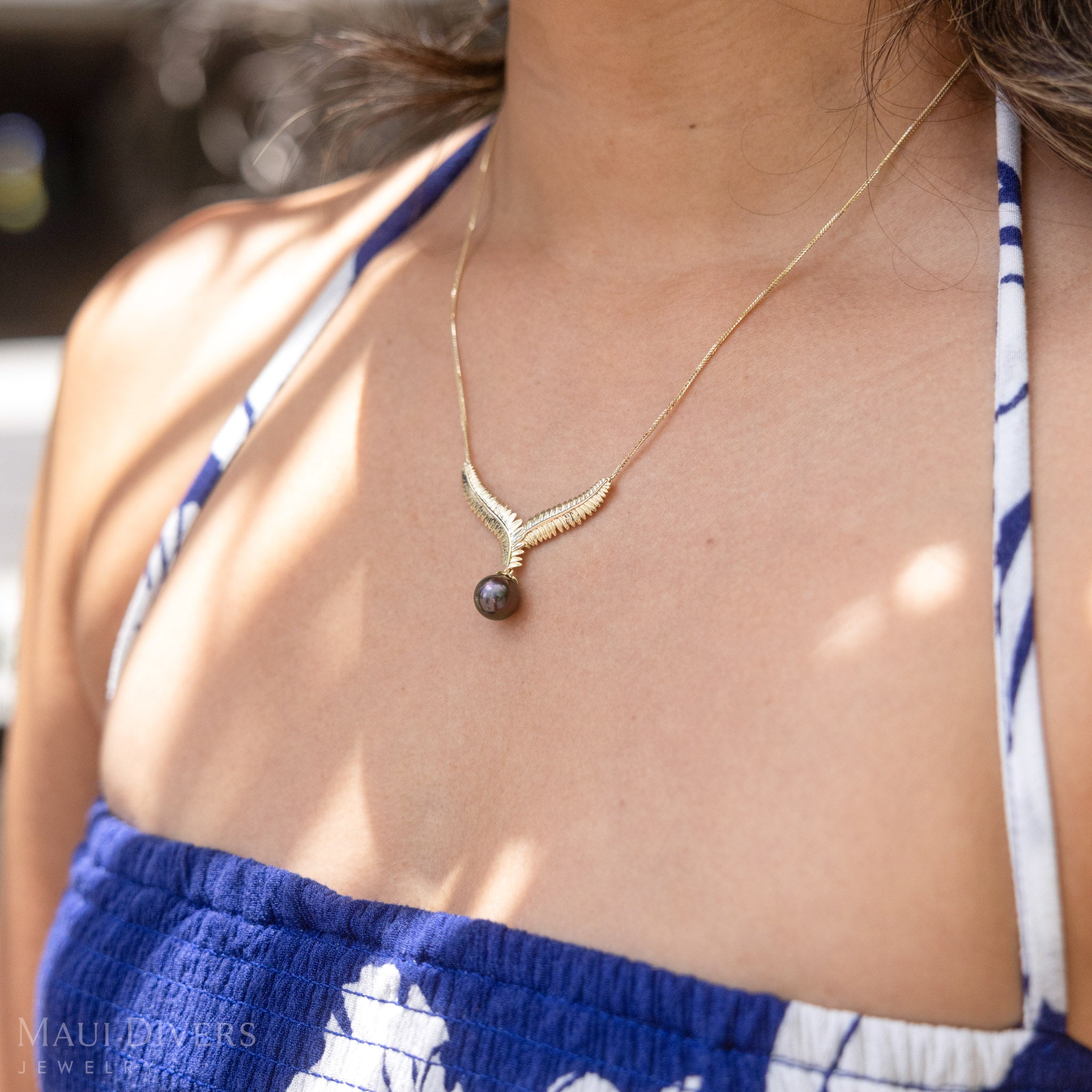 Close-up of a gold kupukupu fern Tahitian black pearl necklace on a person wearing a blue and white dress.