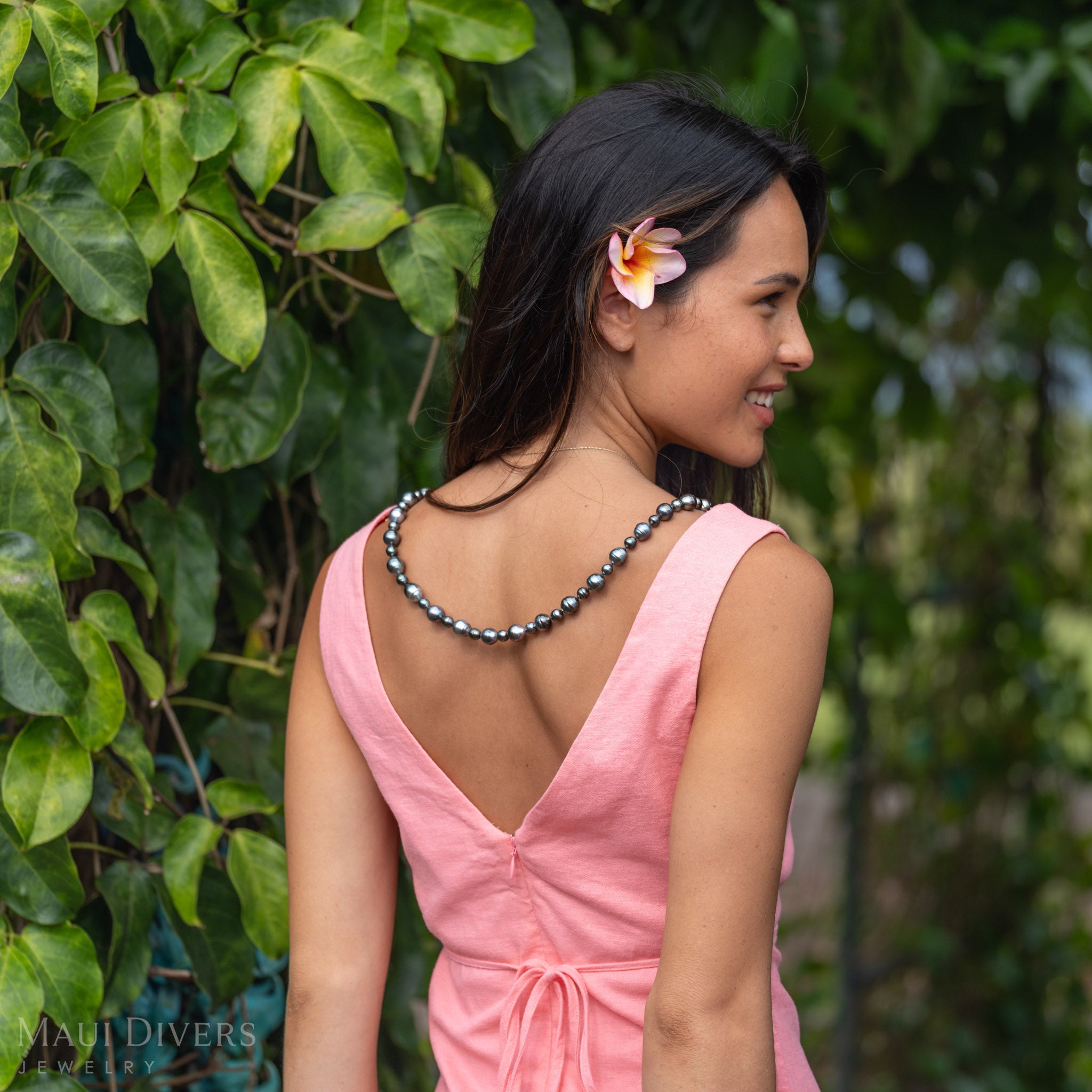 A woman facing away with her head turned back wearing a Natural Tahitian Black Pearl Strand Necklace draped around her back with a pink dress, pink and orange plumeria in her hair, and standing against a green leafy background. 