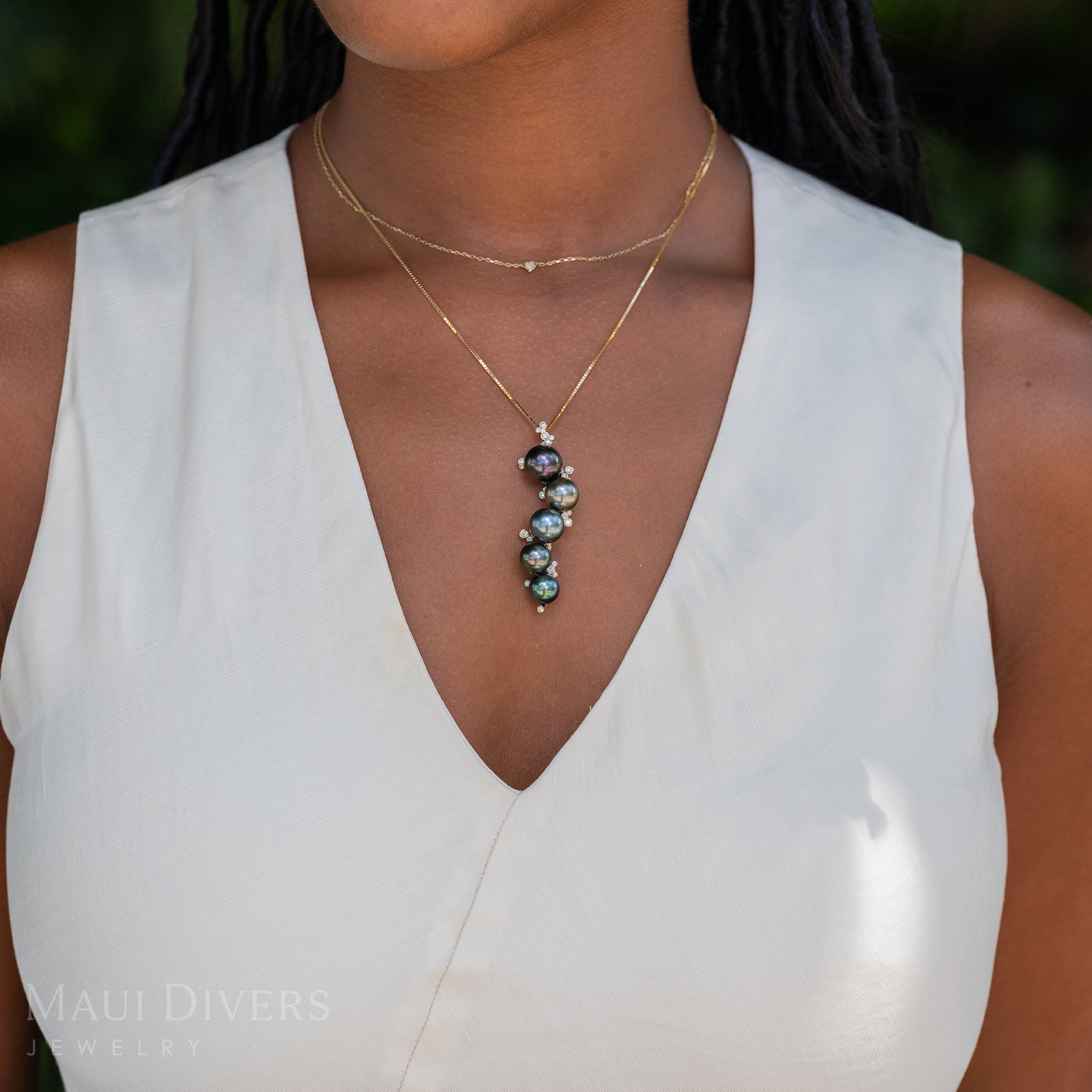 A close-up of a smiling woman in a white top wearing a Bubbling Tahitian Black Pearl Pendant in 14k yellow gold with diamonds on a box chain, layered with a tiny diamond heart necklace, against a blurred leafy green background