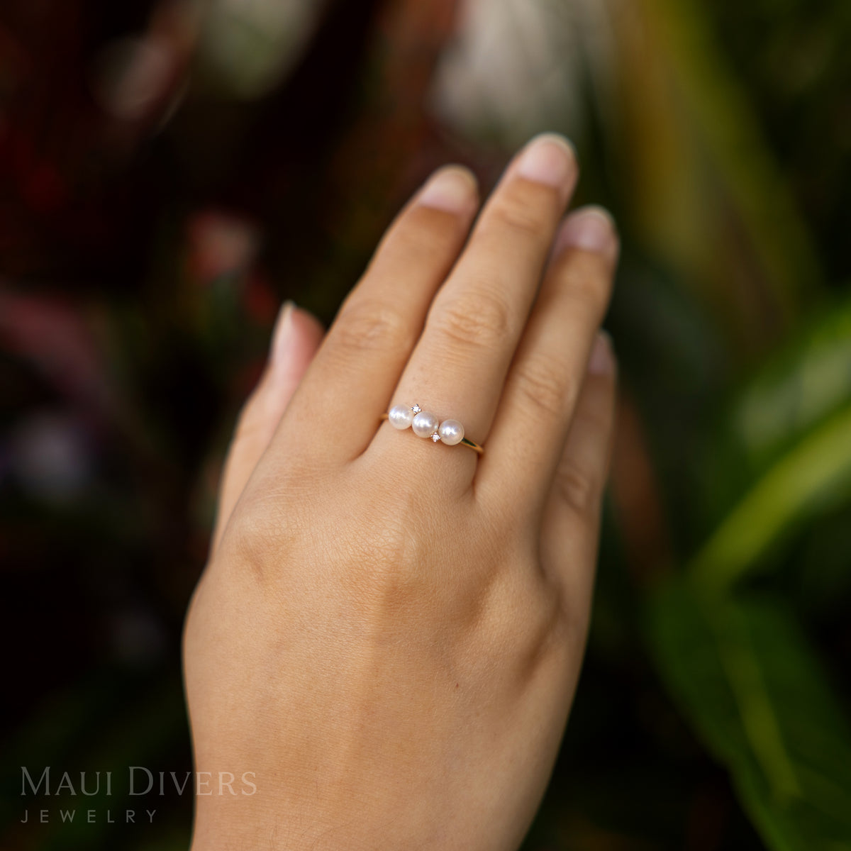 Close-up of a hand wearing an Akoya White Pearl Ring in 14k yellow gold with diamonds, against a blurred red and green leafy background