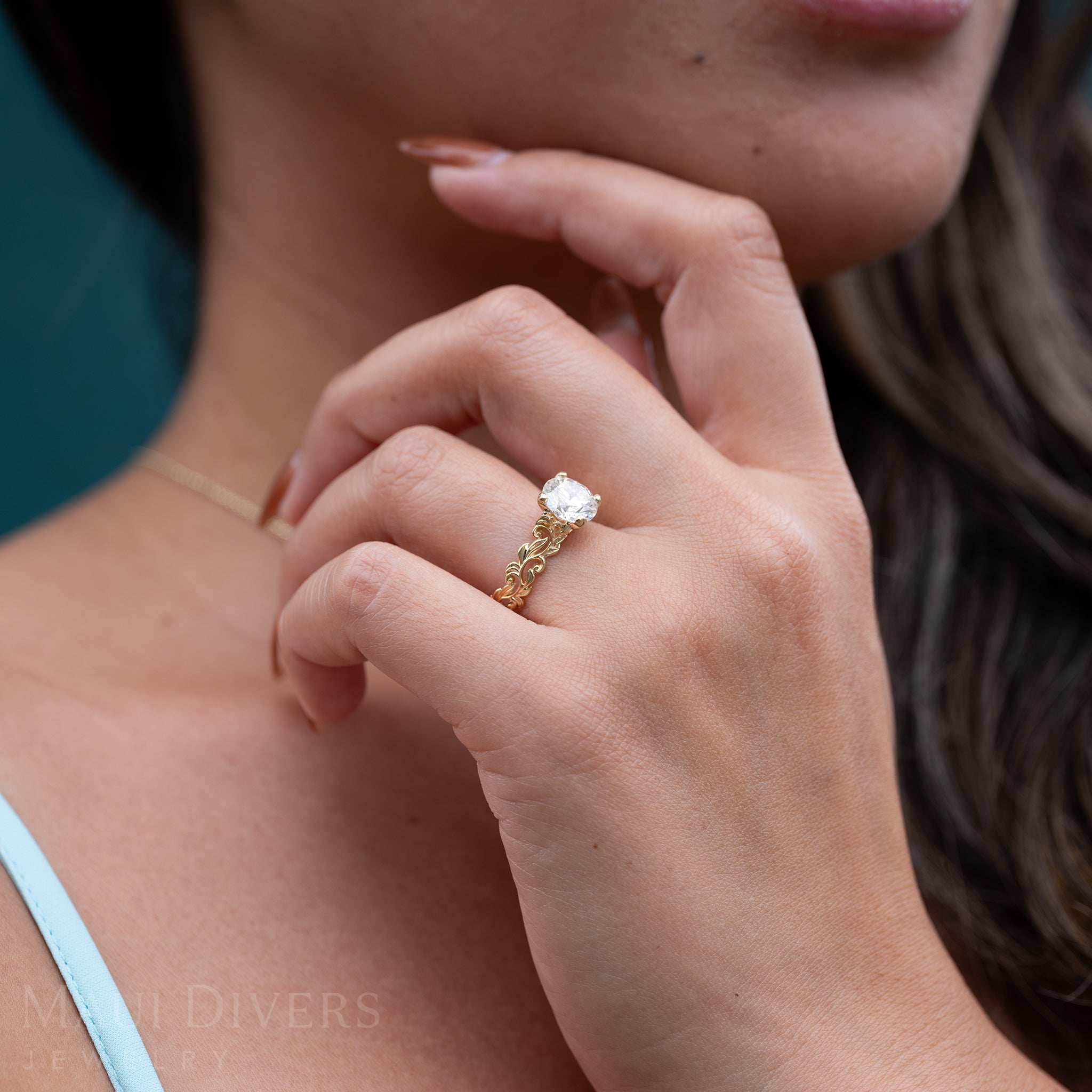 Close-up of a Woman in a Blue Top Wearing A Living Heirloom Engagement Ring in 14k Yellow Gold with a Lab Grown Diamond against a blurred darker blue background