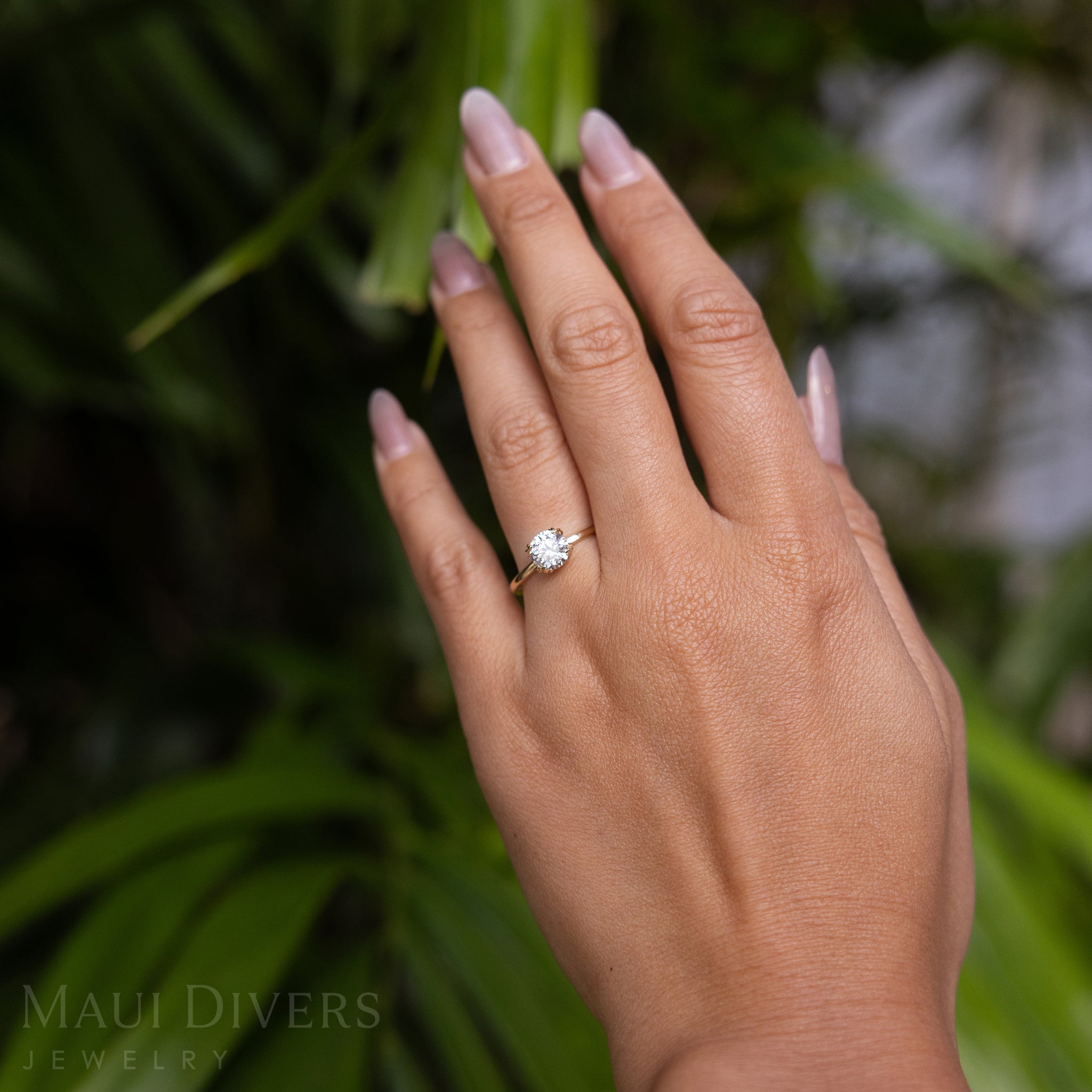 A Woman's Hand Wearing a Hidden Hawaiʻi Maile Ring in 14k Yellow Gold with a Lab Grown Diamond Against a Blurred Green Leaf Background
