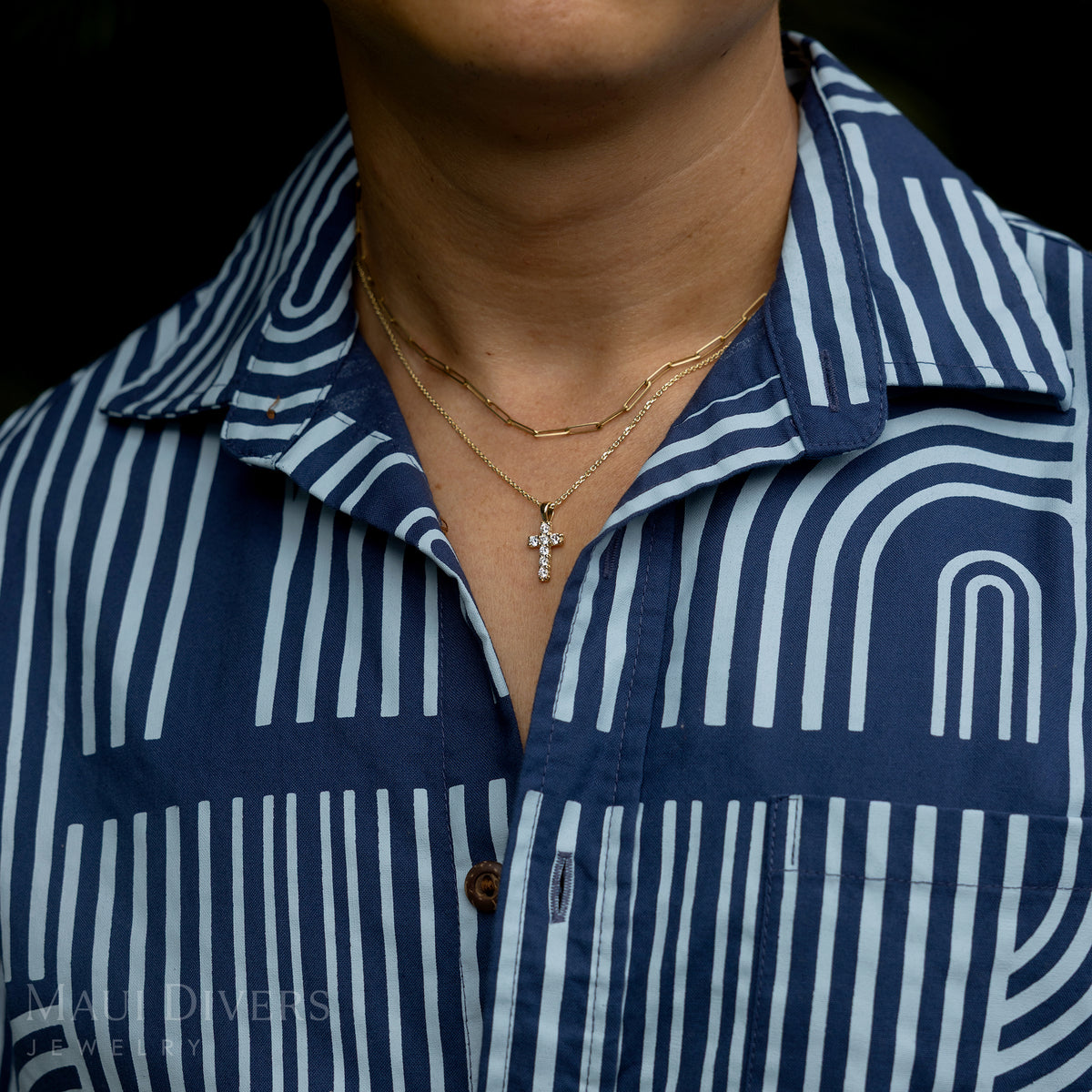 A close-up of a guy in a two-toned blue patterned shirt wearing a Hidden Hawaiʻi Maile Lab Grown Diamond Cross Pendant in 14k yellow gold around his neck, layered with a paperclip chain against a blurred green leaf background