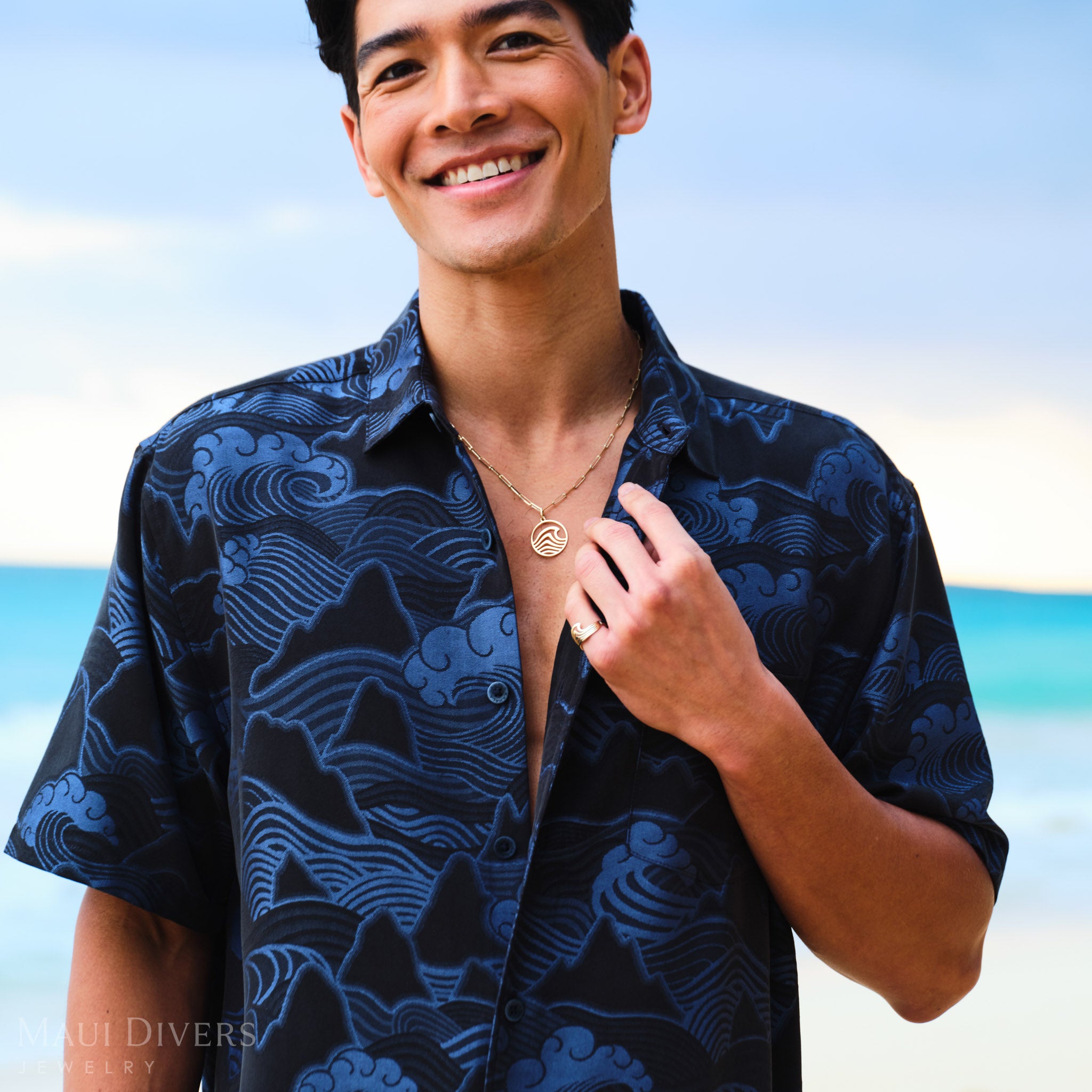 Man wearing a blue patterned shirt with an Abstract Nalu Ring and Pendant in Gold, against a blurred beach background.