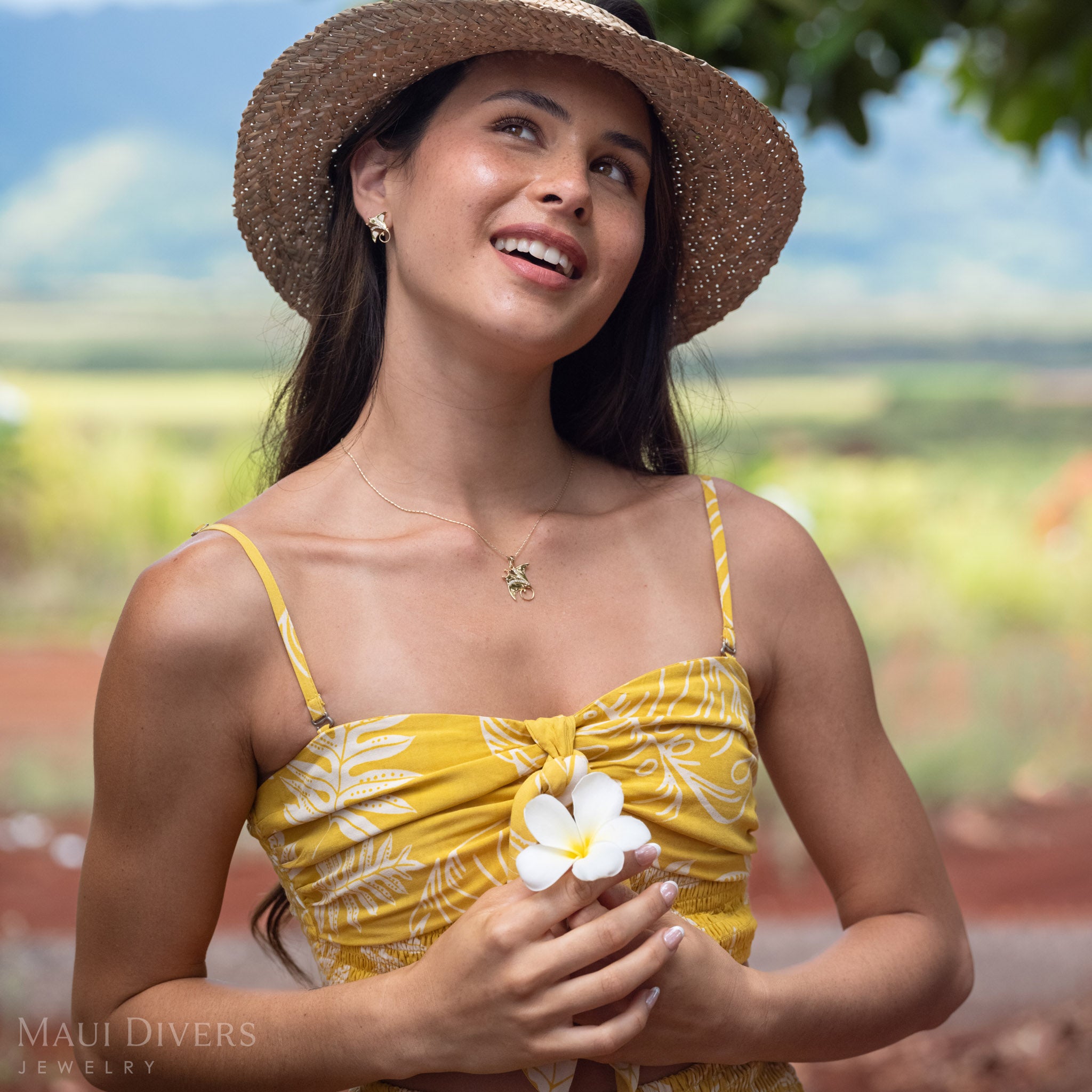 Woman looking up wearing a 14k Gold Manta Ray Earring with Natural Diamond detailing and a matching necklace, holding a white plumeria flower and wearing a brown woven hat. 