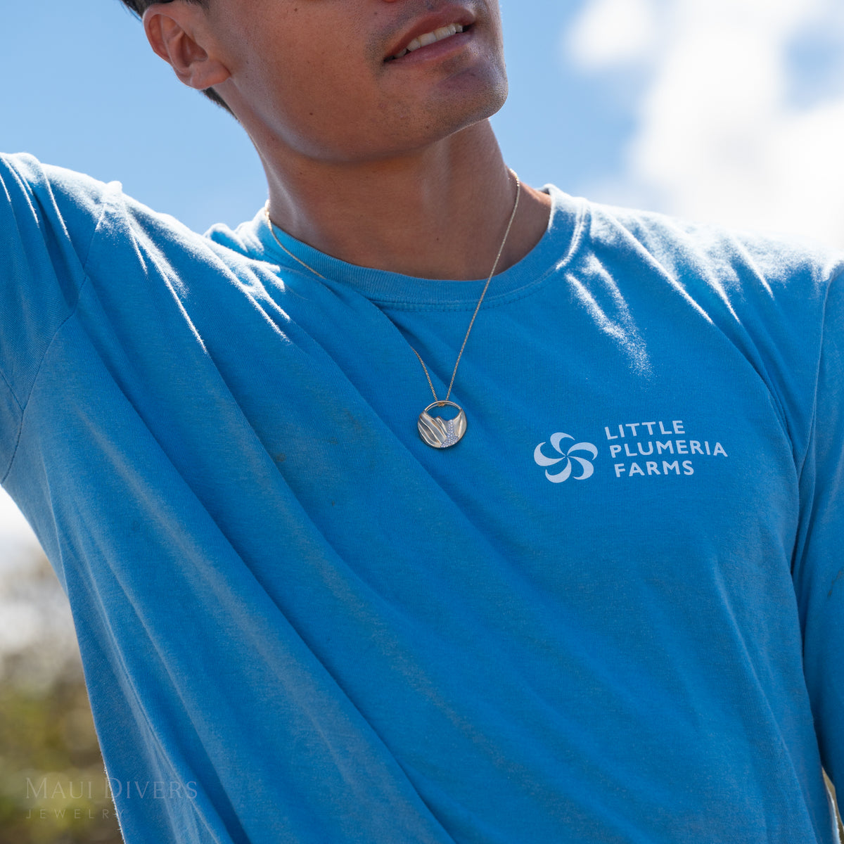 Close up of Dane Little in a blue shirt wearing a round pendant with the koʻolau mountain range with diamonds, against a blurry blue sky