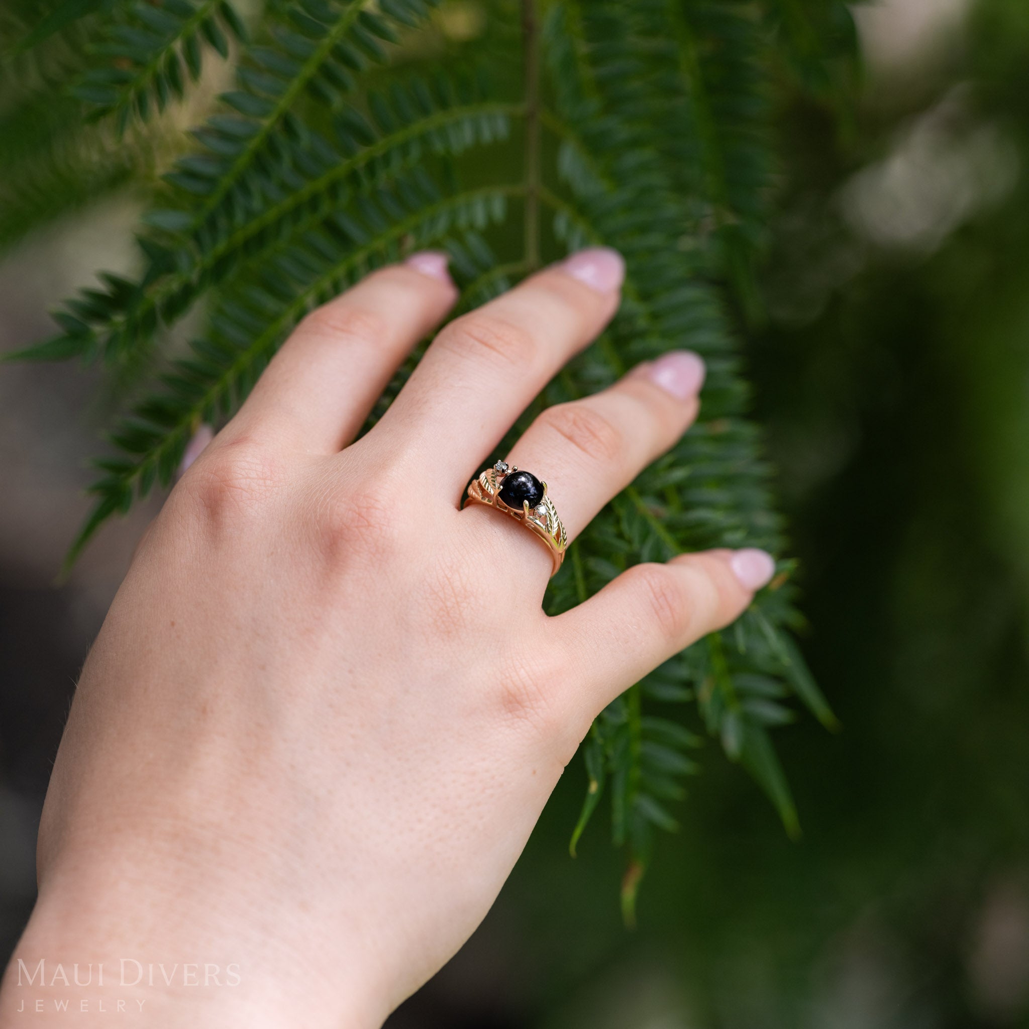 A gold diamond-cut maile leaf ring with Hawaiian black coral and diamonds worn on a hand, positioned against a background of green leaves.