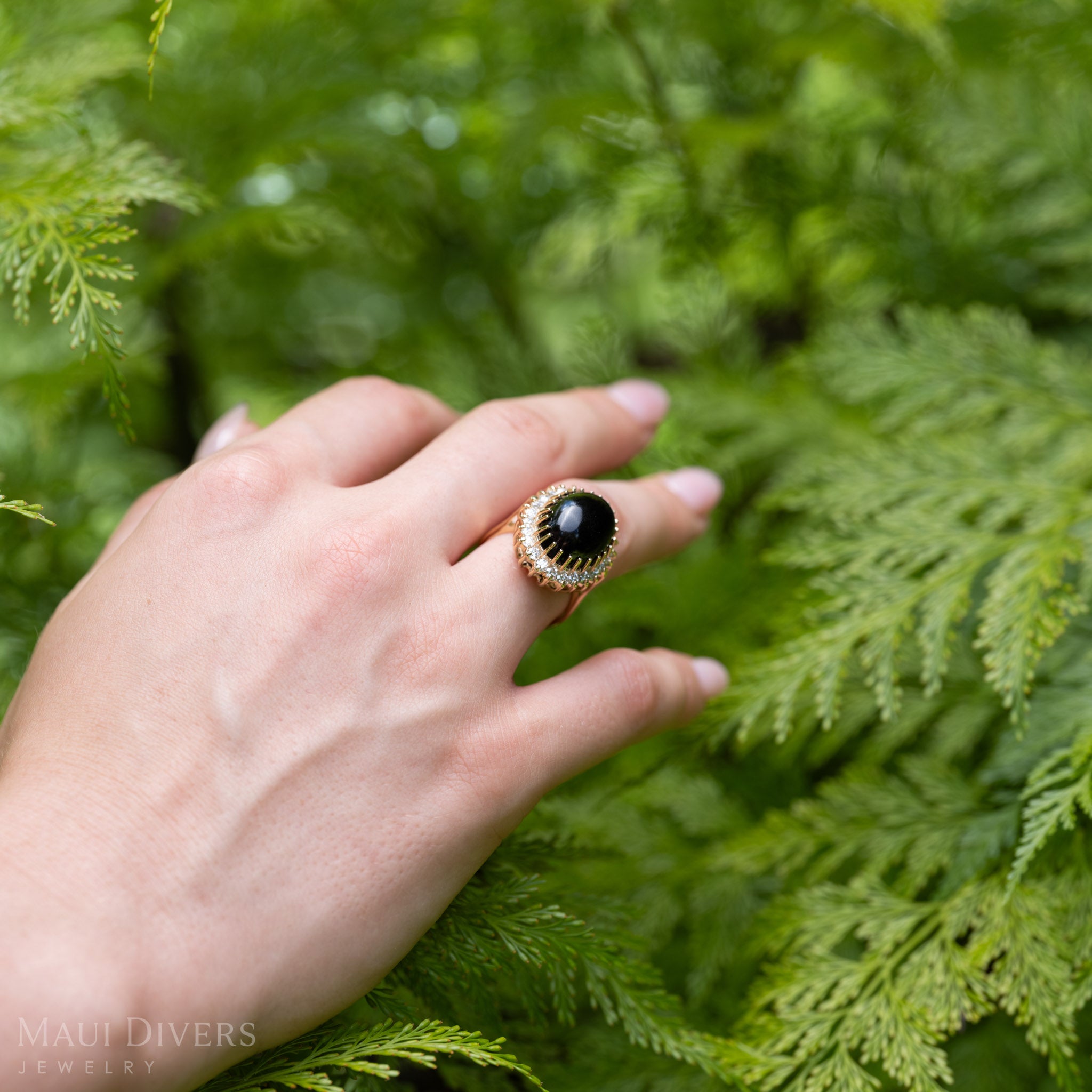 A hand wearing a yellow gold ring with a large oval Hawaiian black coral cabochon and a halo of diamonds, set against a background of green fern foliage. 