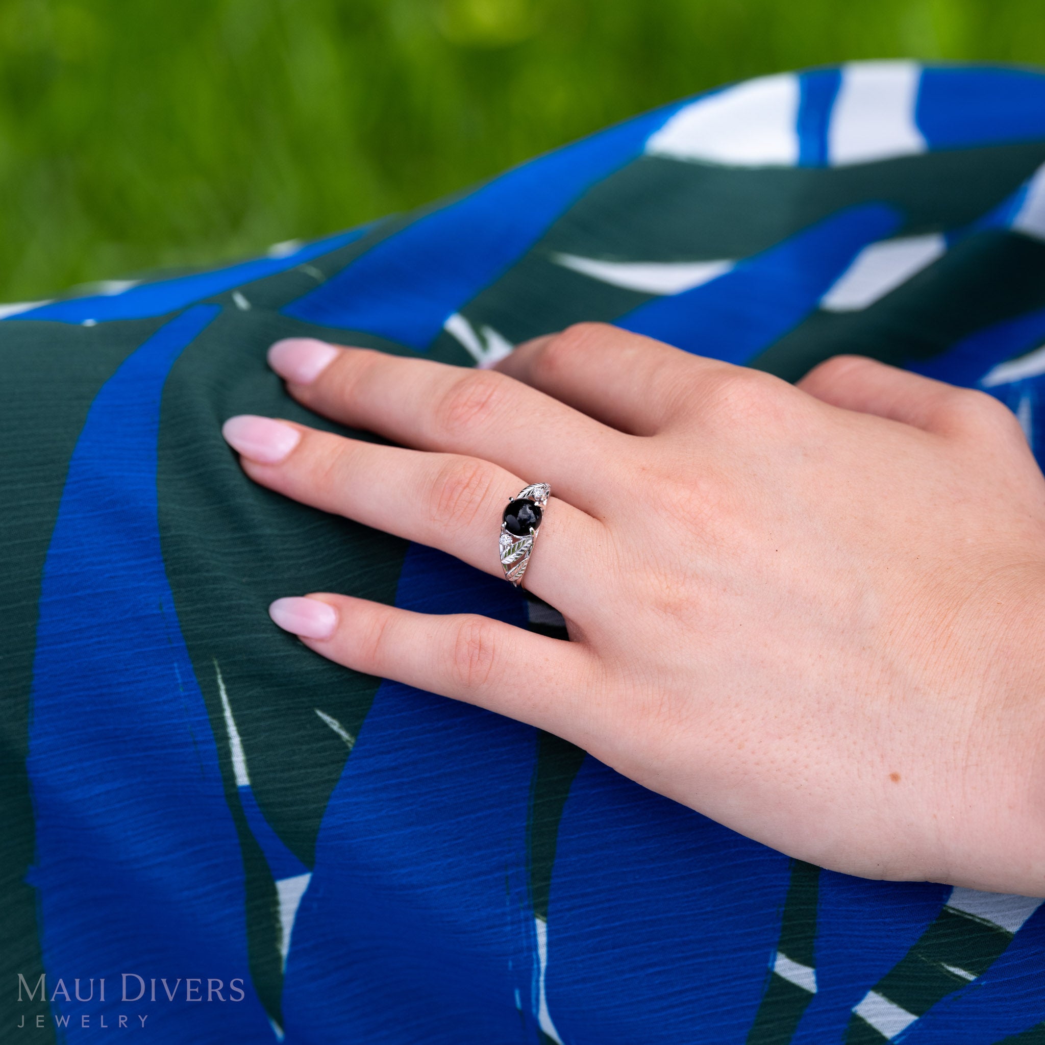 A close-up of a person's hands resting on a blue and green patterned garment, wearing white gold ring with diamond-cut maile leaves, diamond accents, and Hawaiian black coral. The background is a soft-focus green outdoor setting.