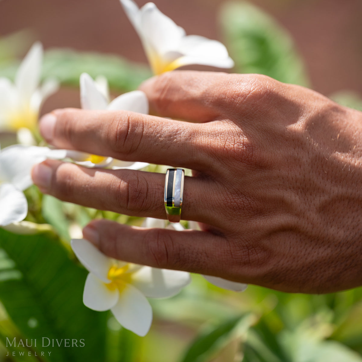 Close-up of a hand reaching for plumeria flowers and wearing a gold ring with horizontal inlay of black coral and mother of pearl