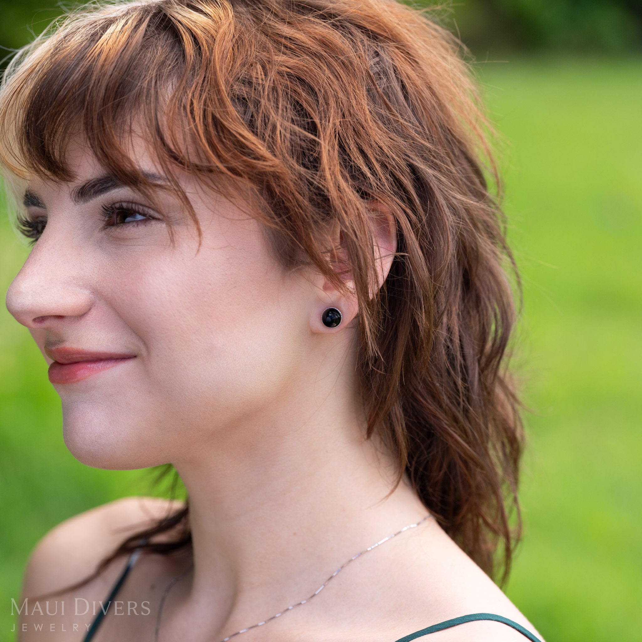 A close-up side profile of a woman with wavy light brown hair wearing a diamond halo Hawaiian black coral bead stud earring, set against a blurred green outdoor background. 