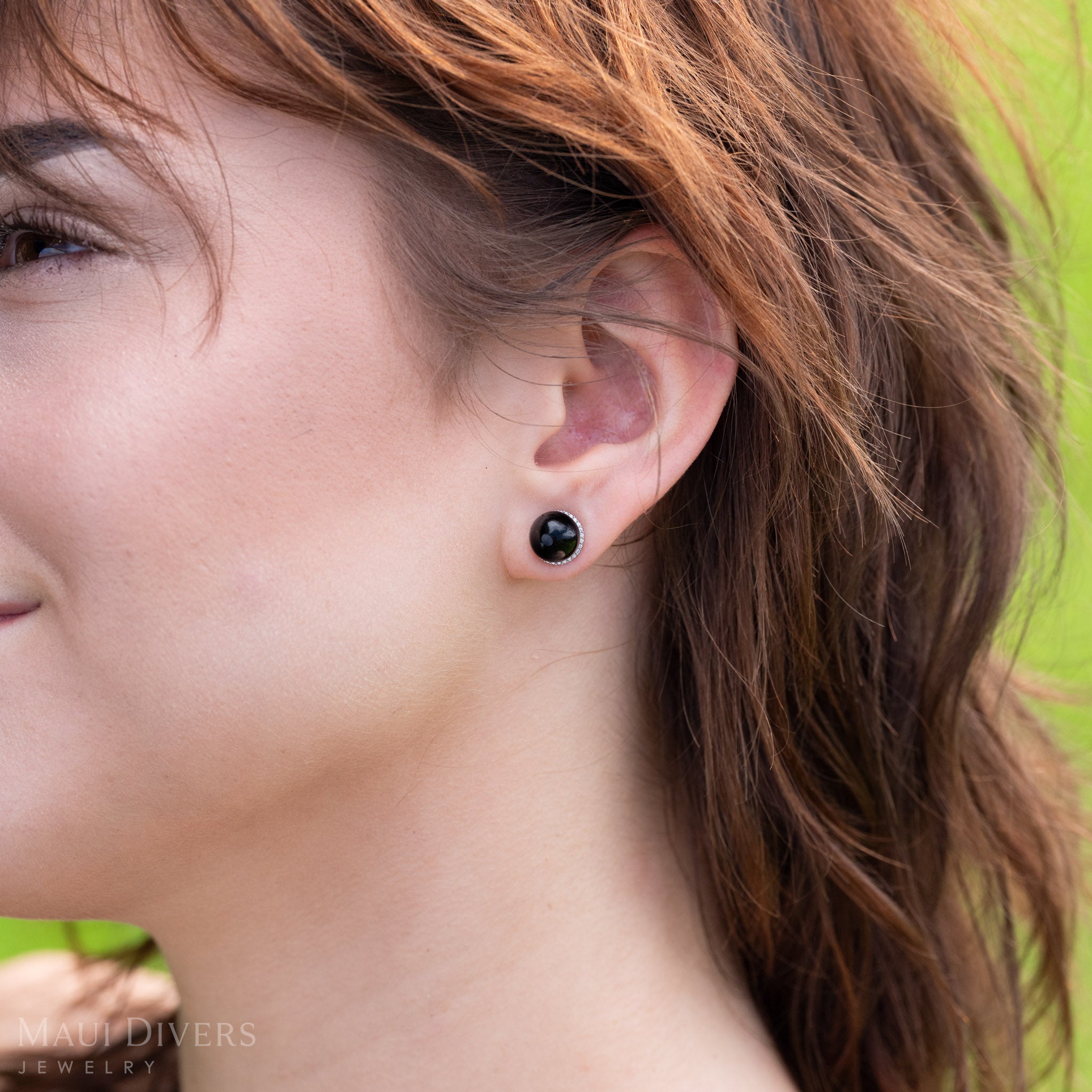 A close-up side profile of a woman wearing diamond halo Hawaiian black coral bead earring in white gold on her left ear against a blurred green outdoor background. 