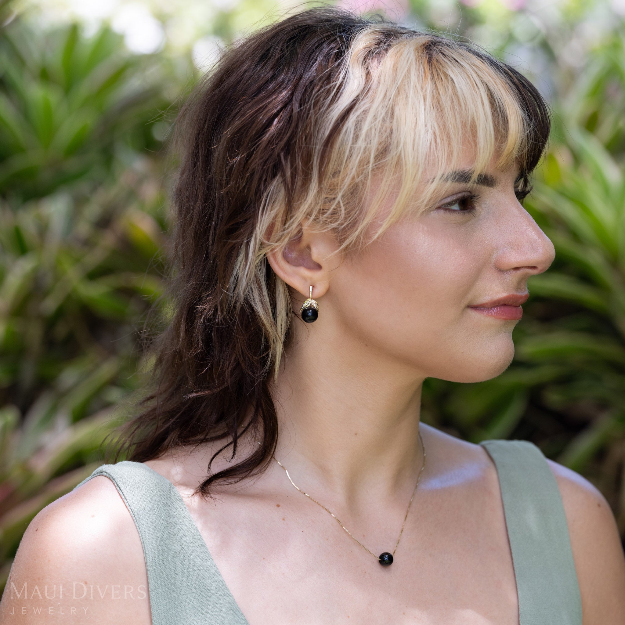 A woman wearing a diamond-cut maile leaf and Hawaiian black coral bead stud earring, photographed outdoors with green foliage in the background.
