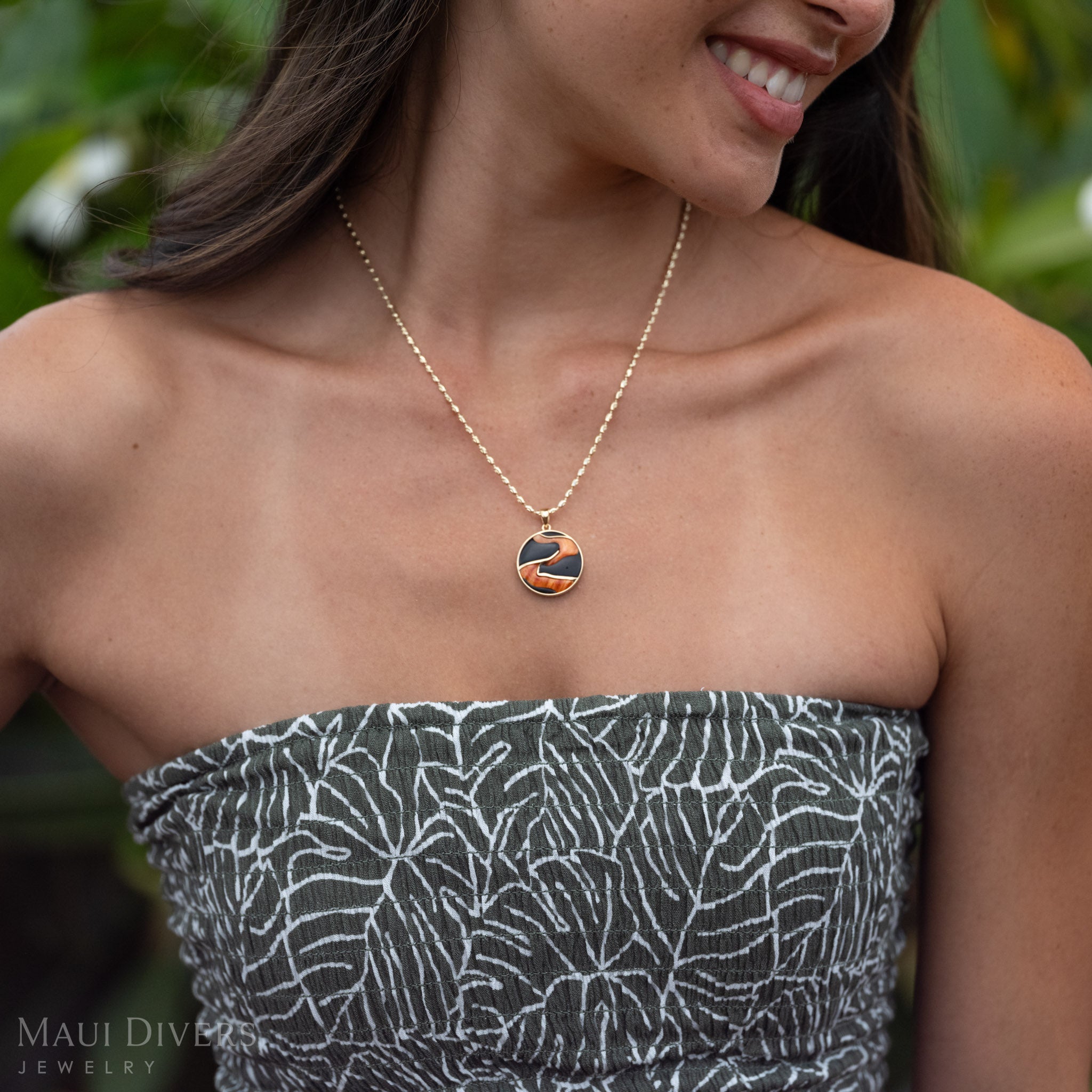 Close-up of a woman smiling wearing Lava Flow Black Coral and Spiny Oyster Pendant in 14k Yellow Gold in front of a blurred leafy background