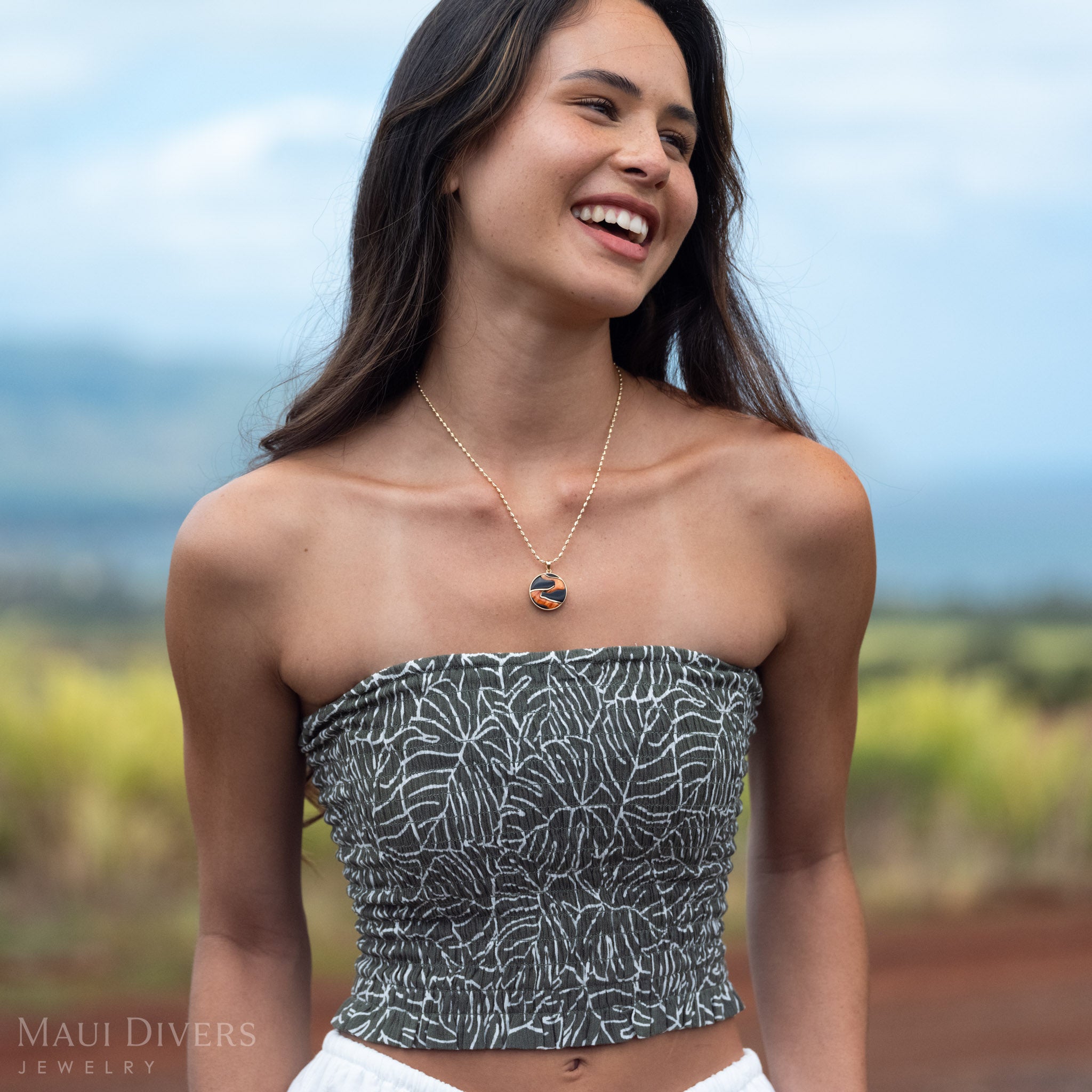 Woman smiling wearing Lava Flow Black Coral and Spiny Oyster Pendant in 14k Gold in front of a blurred mountain landscape. 
