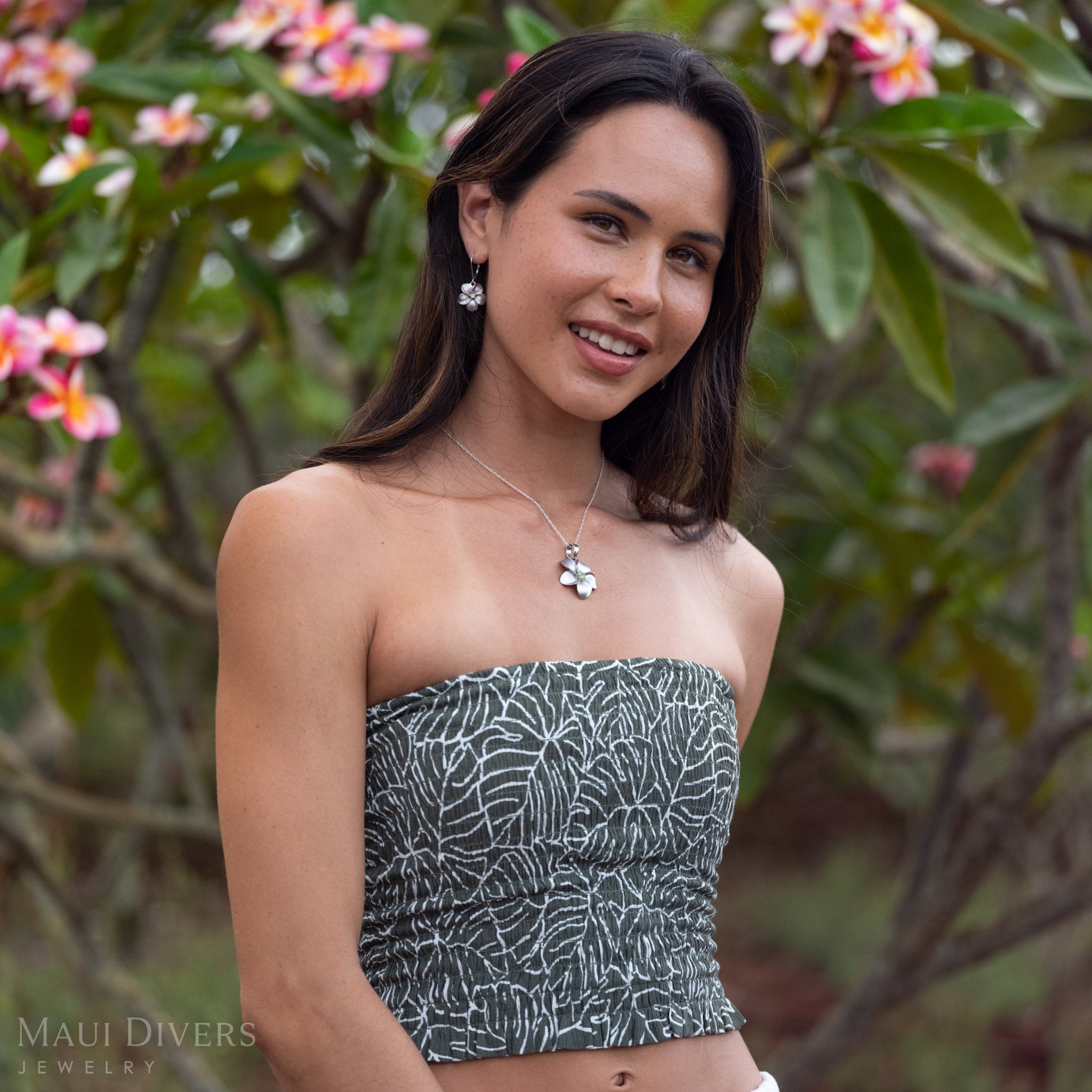 Woman wearing Peridot Plumeria Earrings in Sterling Silver with a necklace in front of a blurred plumeria tree with green leaves and pink flowers. 
