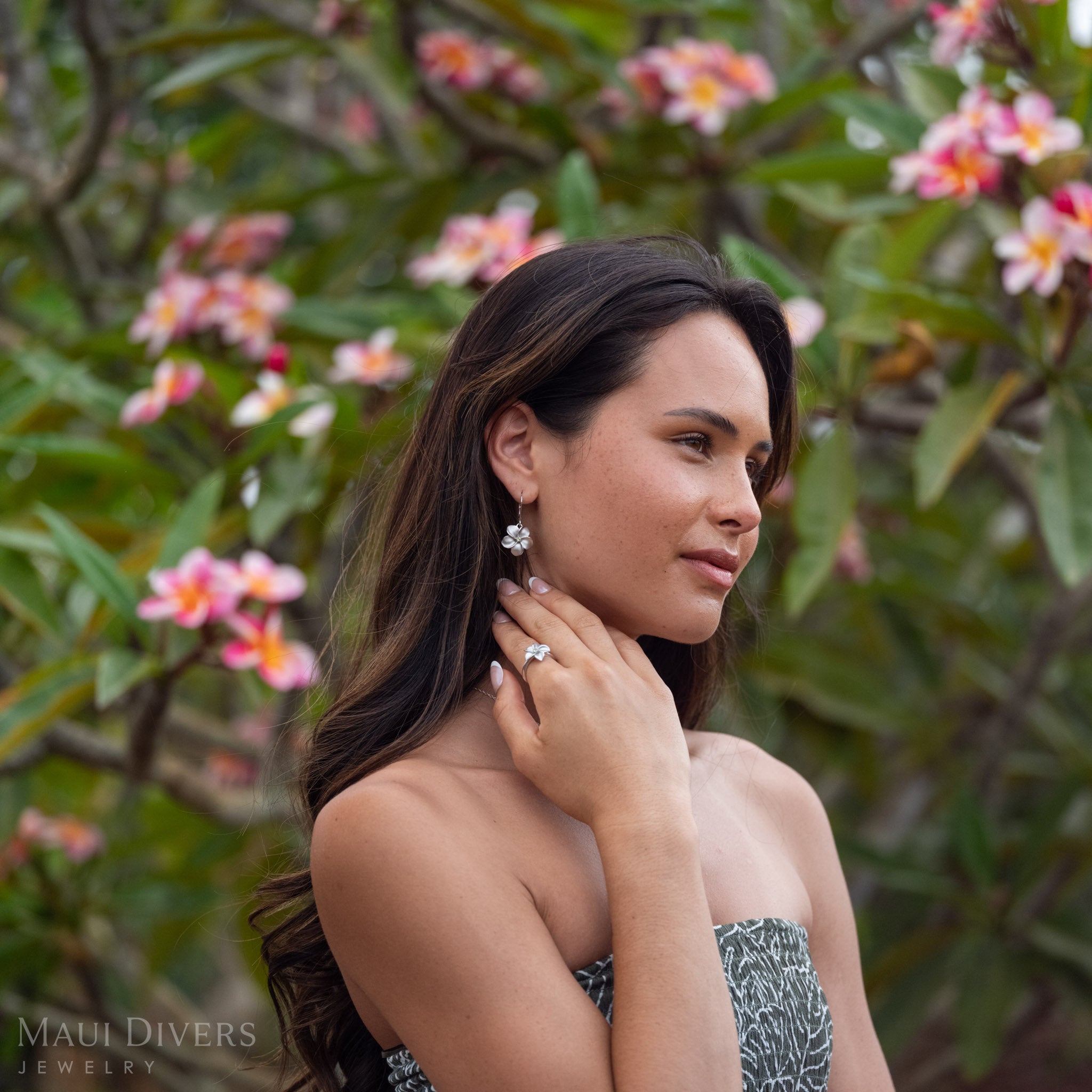 Woman gazing to the side wearing Peridot Plumeria Earrings in Sterling Silver with a matching ring in front of a blurred plumeria tree with green leaves and pink flowers. 