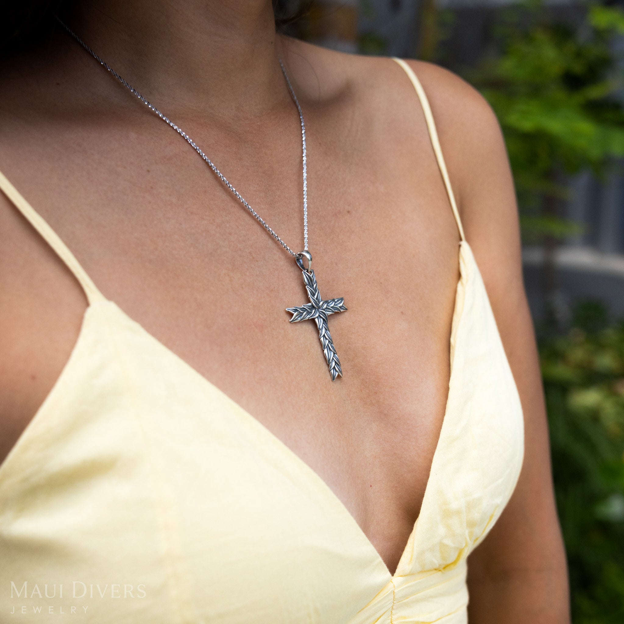 Close-up sideview of a woman in a yellow top wearing a Maile Cross Pendant in Sterling Silver against a blurred leafy green background
