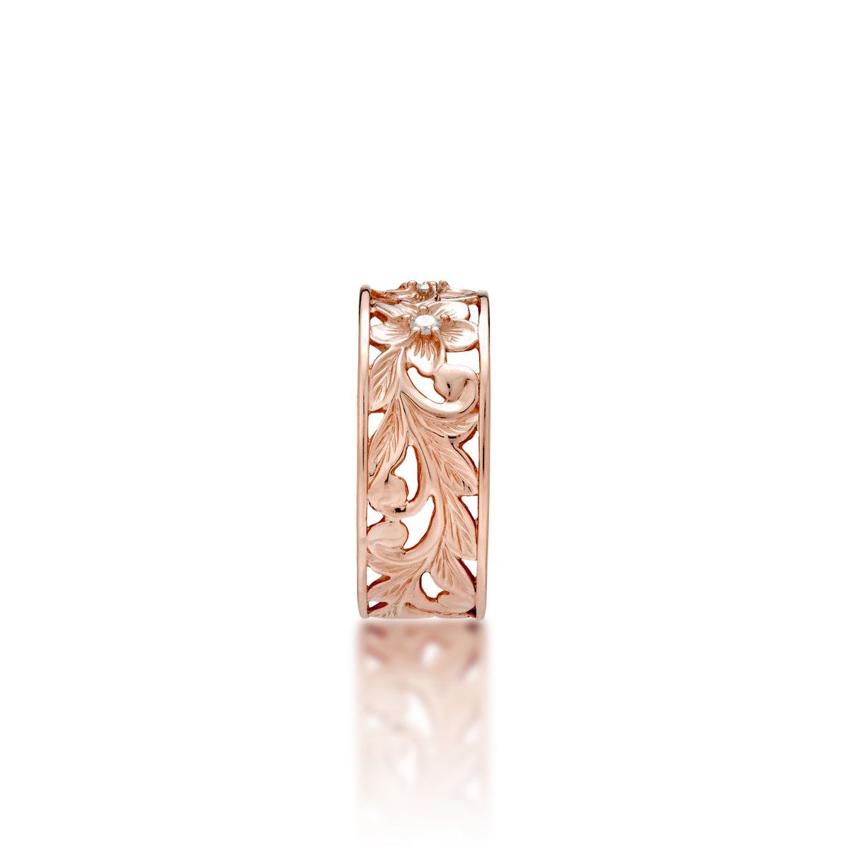 A side view of a 14k rose gold Hawaiian Heirloom ring with an intricate fern and maile leaf scrollwork pattern featuring plumeria flowers at the top with diamond accents, shown on a clean white background with a soft reflection.