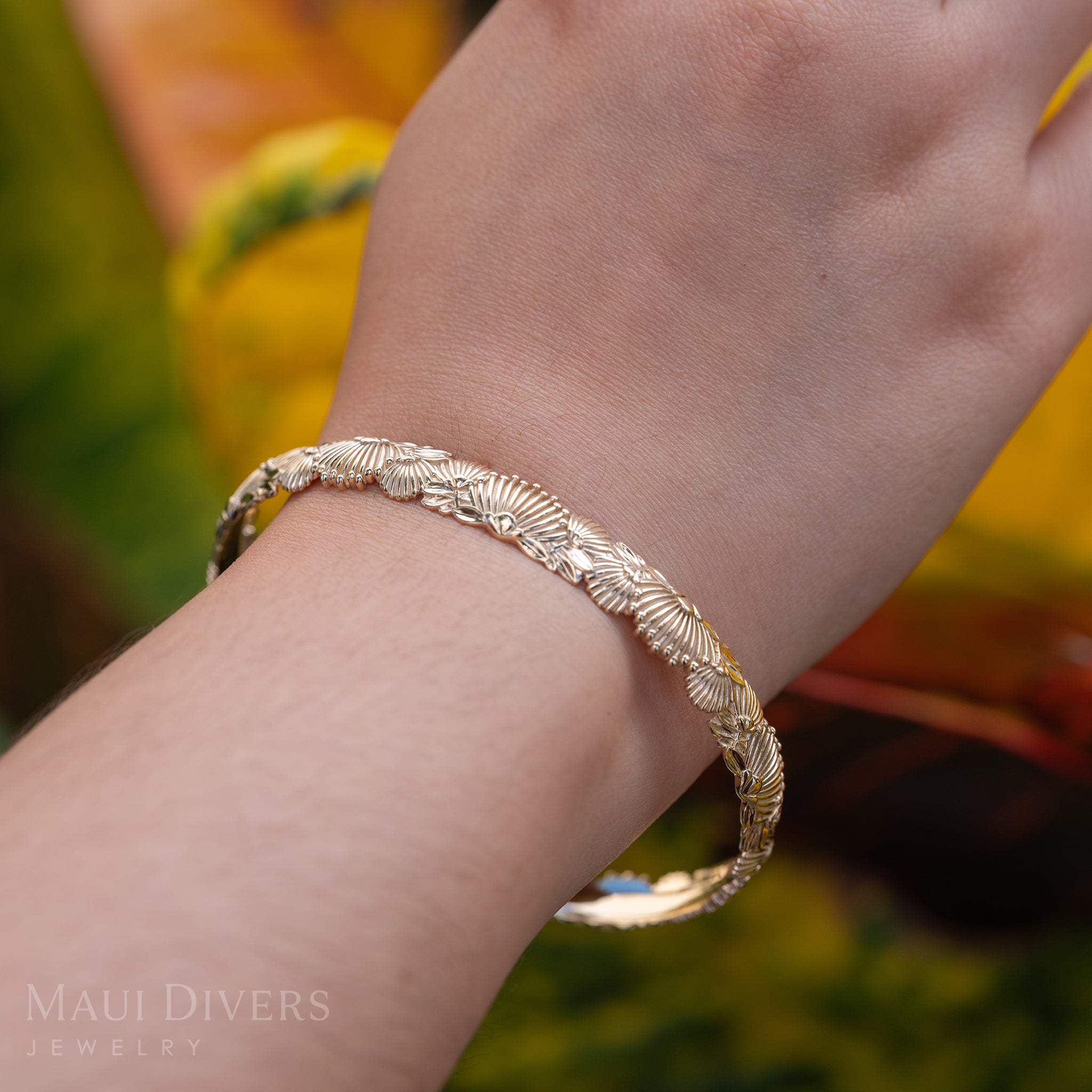 A close-up of an ʻŌhiʻa Lehua Bracelet in 14k yellow gold on a wrist, against a blurred yellow and green leafy background