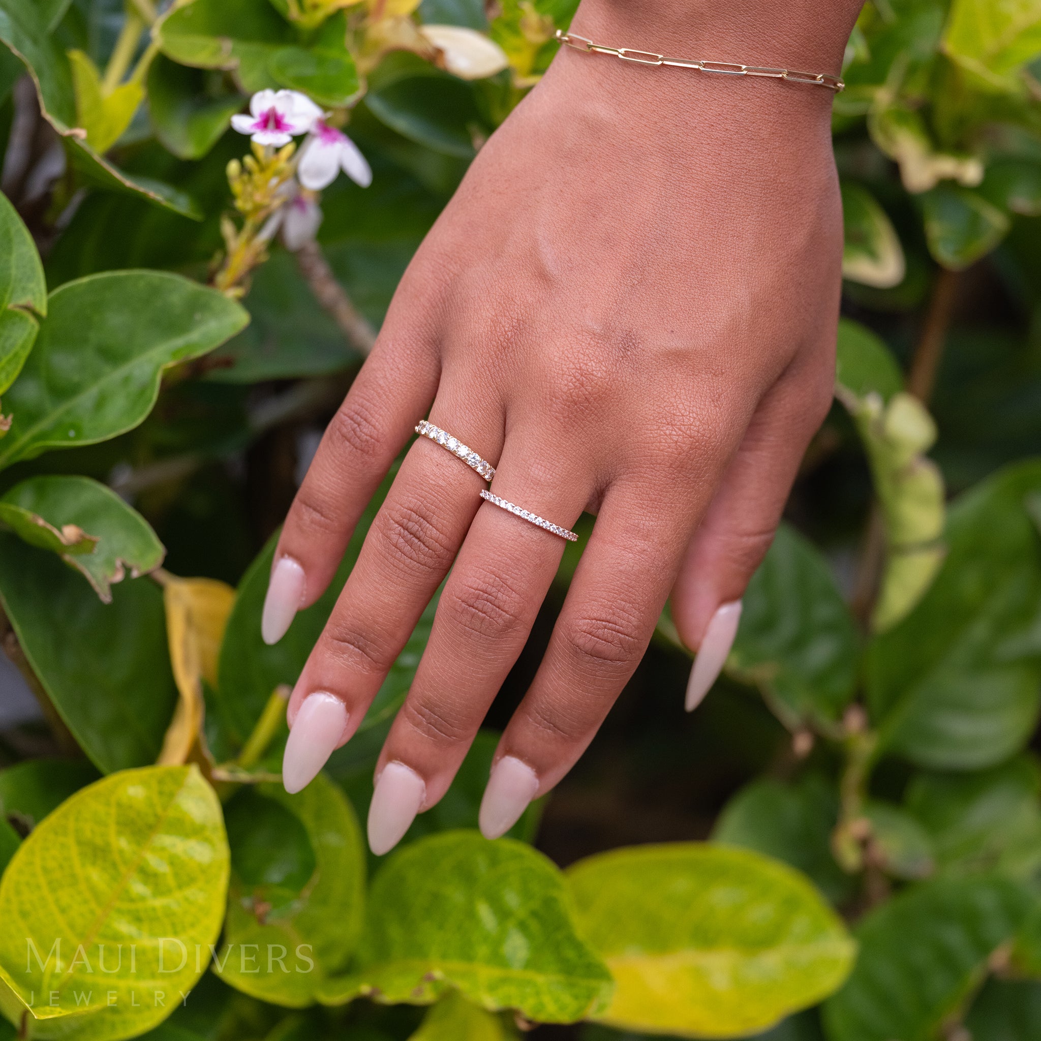 A close-up of a hand wearing an Anniversary Ring 14k rose gold with diamonds, paired with a Hidden Hawaiʻi Maile Anniversary ring in 14k yellow gold with lab grown diamonds, and a paperclip chain bracelet, against a blurred leafy green background