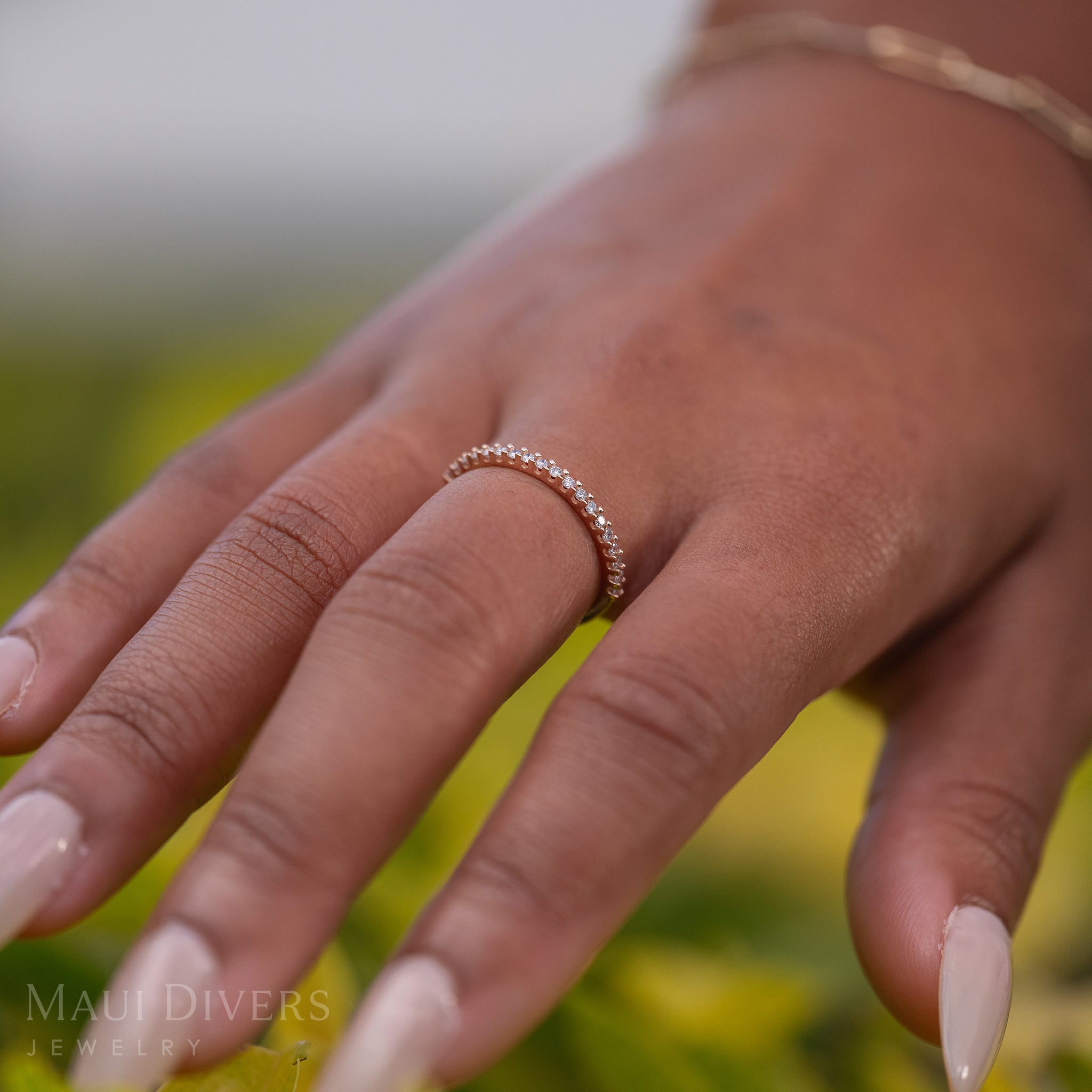 A side-view close-up of a hand wearing an Anniversary Ring in 14k rose gold with diamonds, against a blurred leafy green background