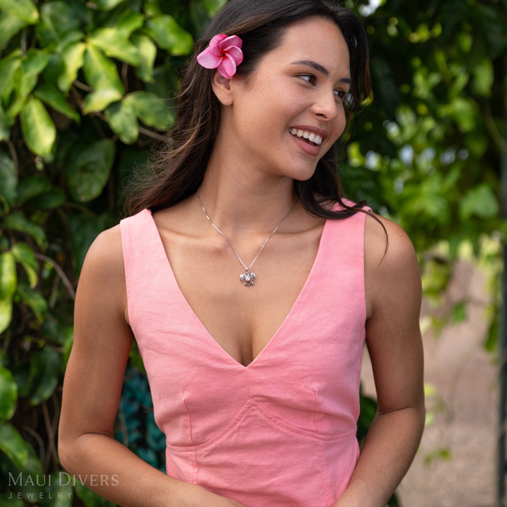 Woman looking to the side wearing a Pick A Pearl Honu Pendant in Sterling Silver with a Natural Pink Freshwater Pearl in front of a green leafy background. 