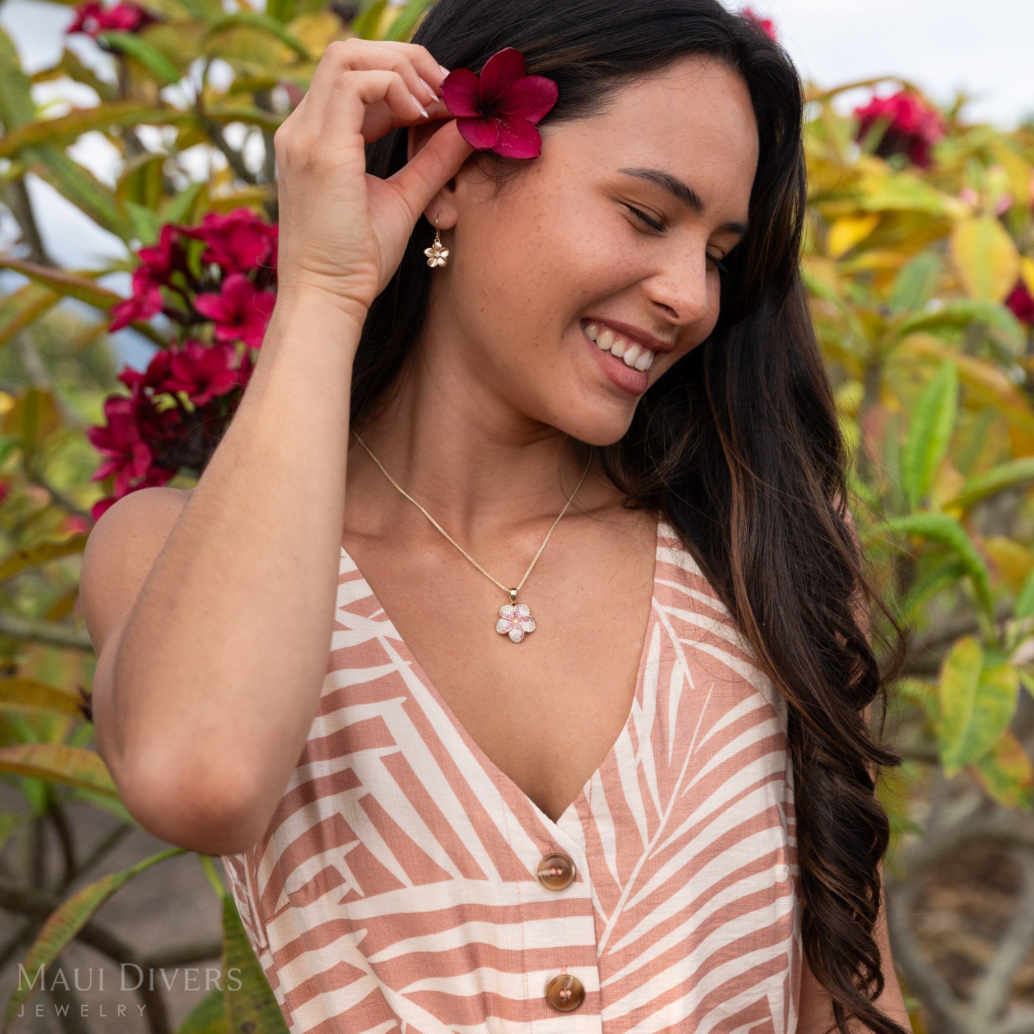 Woman putting a red flower in her hair wearing a 14k Gold Plumeria Earring with Natural Diamond Center and a matching Plumeria Necklace. 